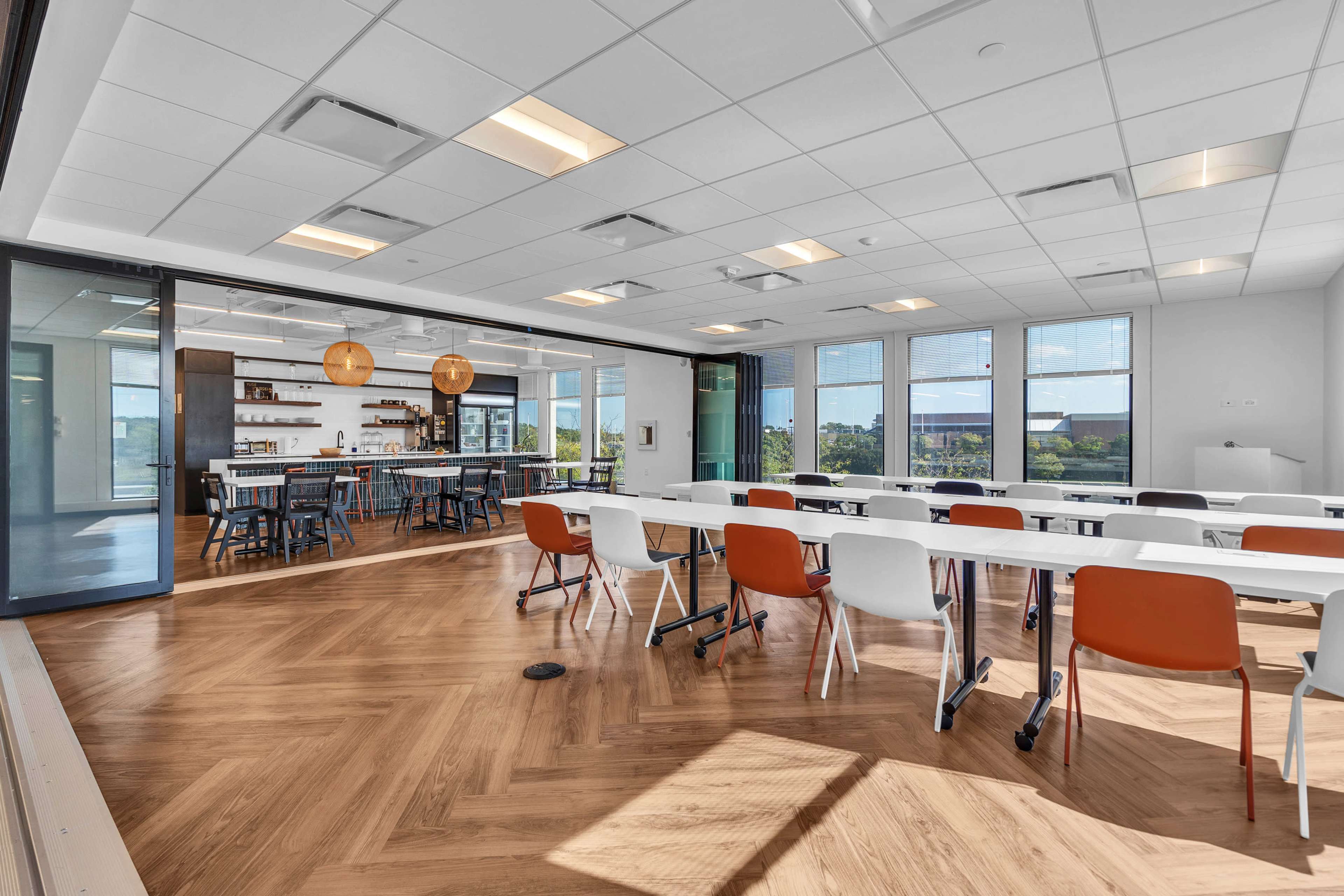 The image shows a modern conference room with white tables and colorful chairs, featuring large windows and an adjoining area with a kitchenette and casual seating.