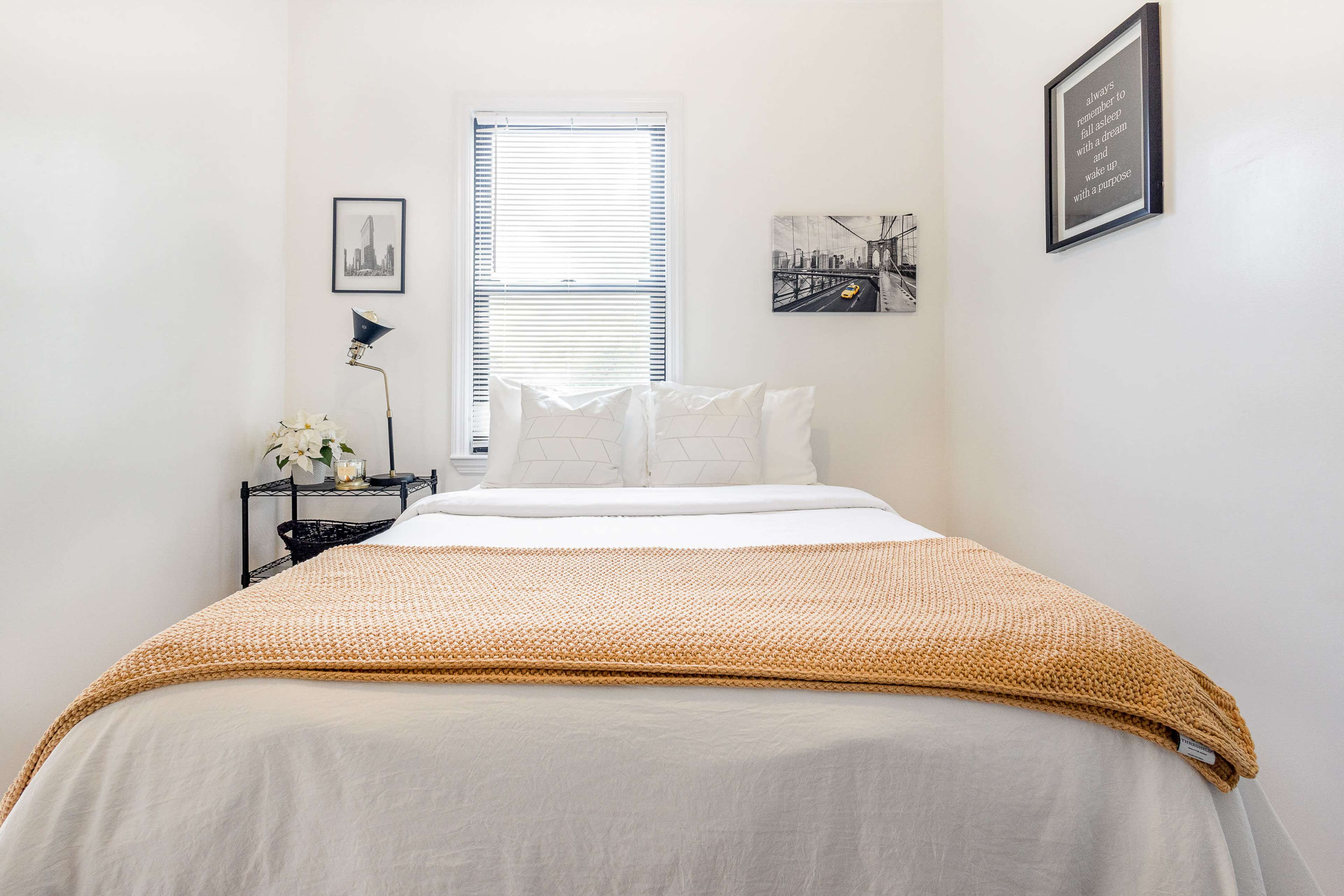 A small, well-lit bedroom with a double bed covered in a white duvet and an orange throw, accompanied by wall art and a window.