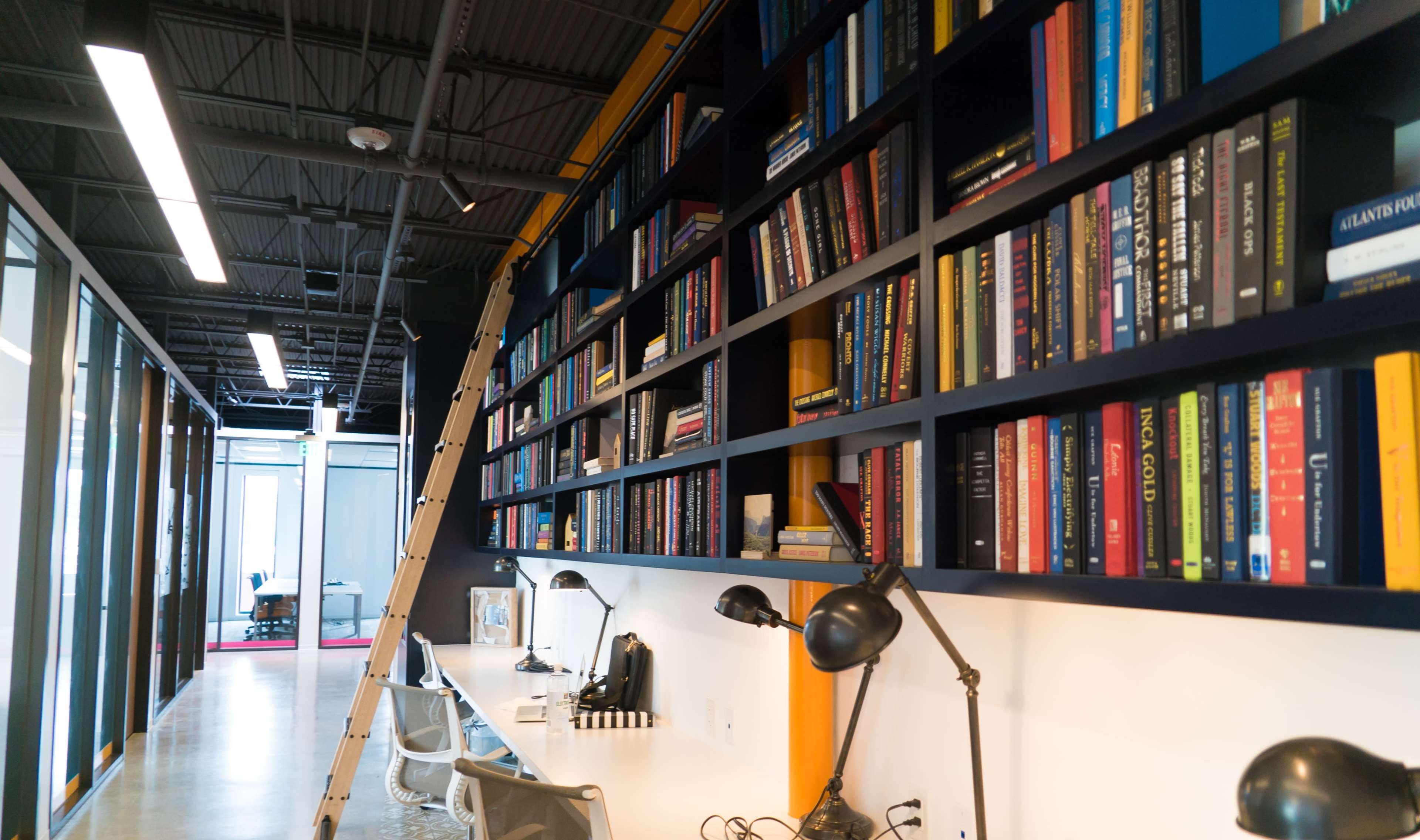 A modern office space features a long, dark shelving unit filled with books, complemented by a white desk area and a metal ladder for access.