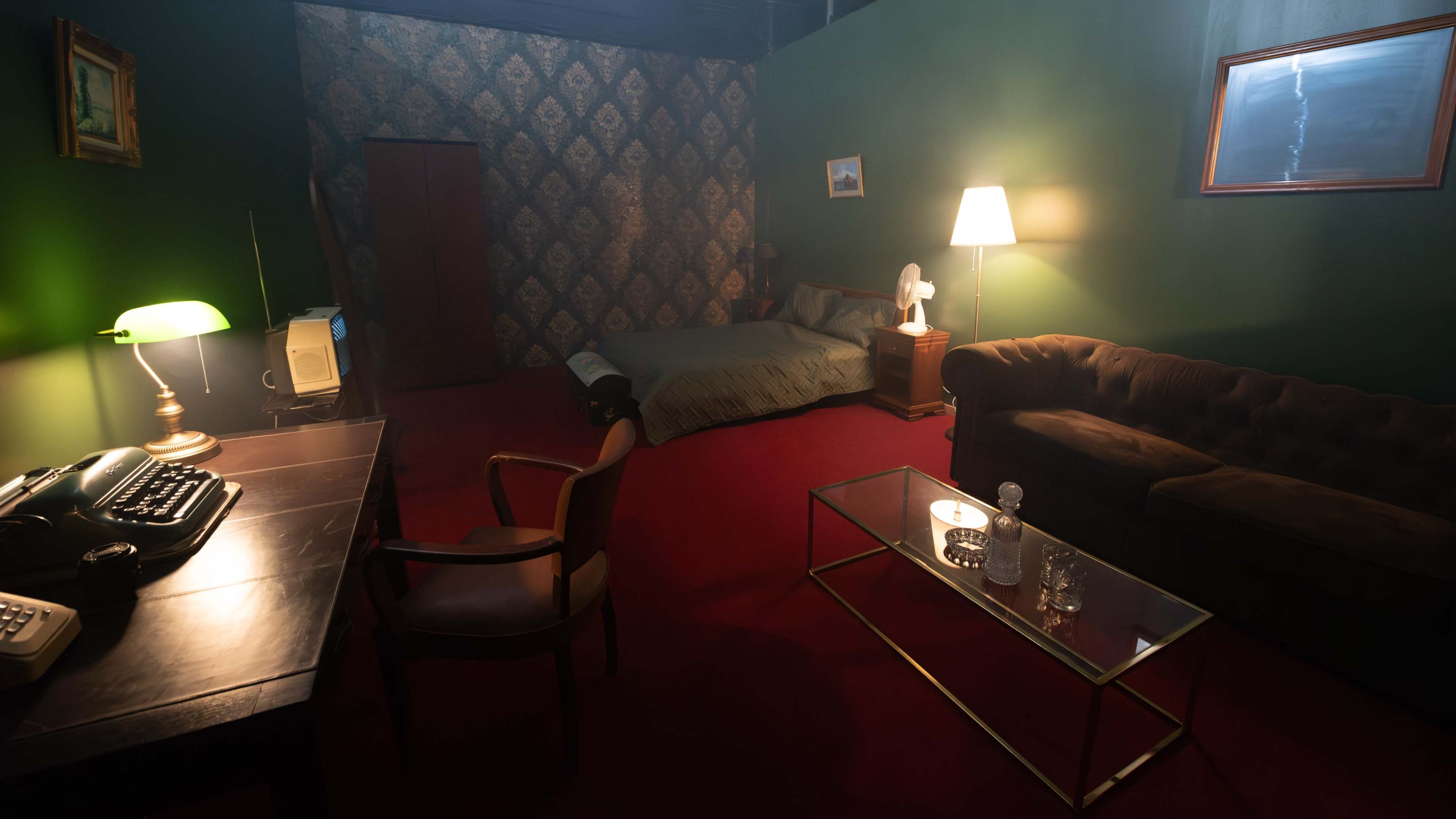 A dimly lit room with a desk, a vintage typewriter, a bed against the wall, a couch, and a lamp on a side table.
