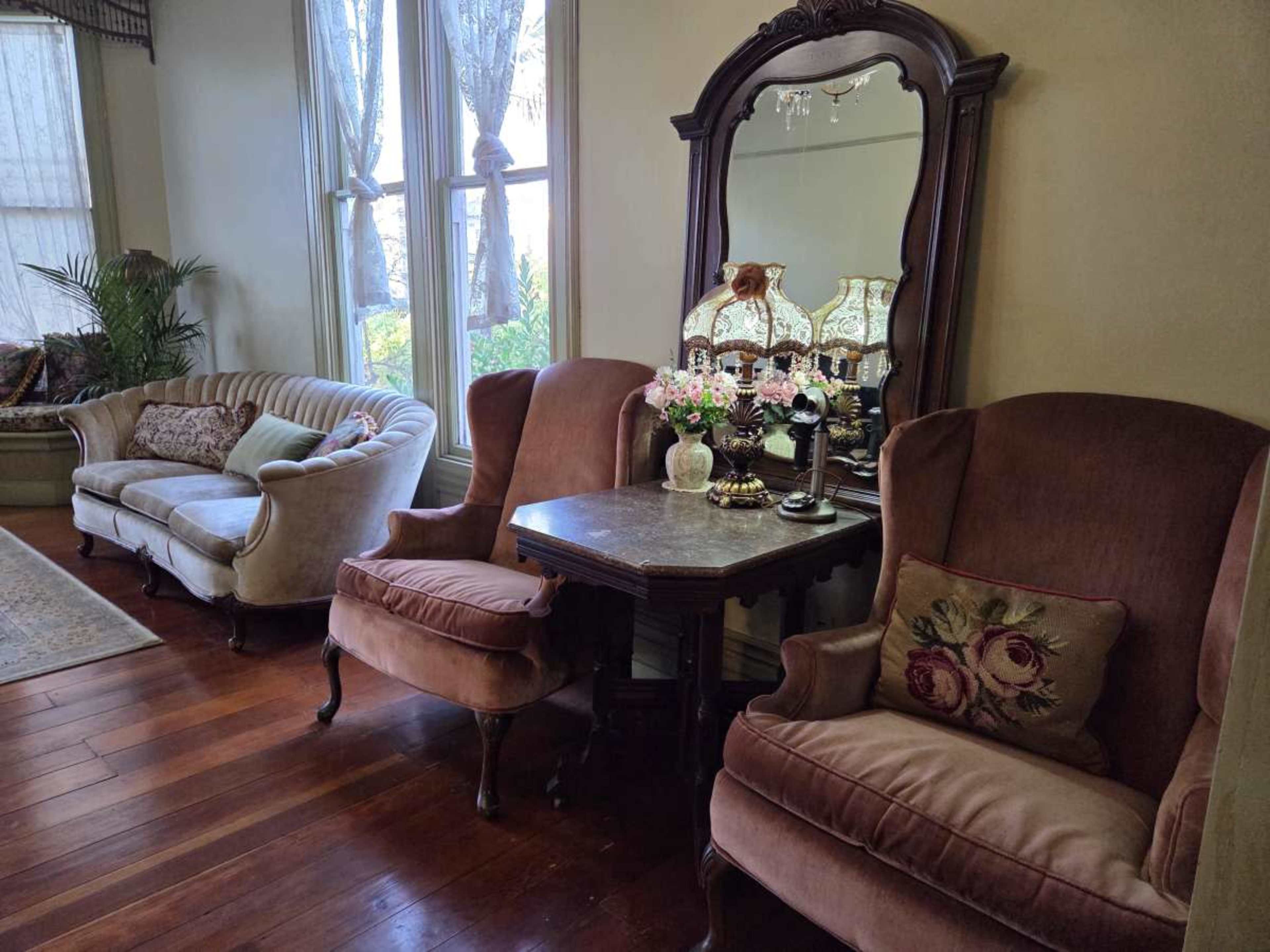 The image shows a vintage sitting area with two upholstered armchairs, a small table with floral arrangements, and a large wall mirror, all set against a backdrop of large windows.
