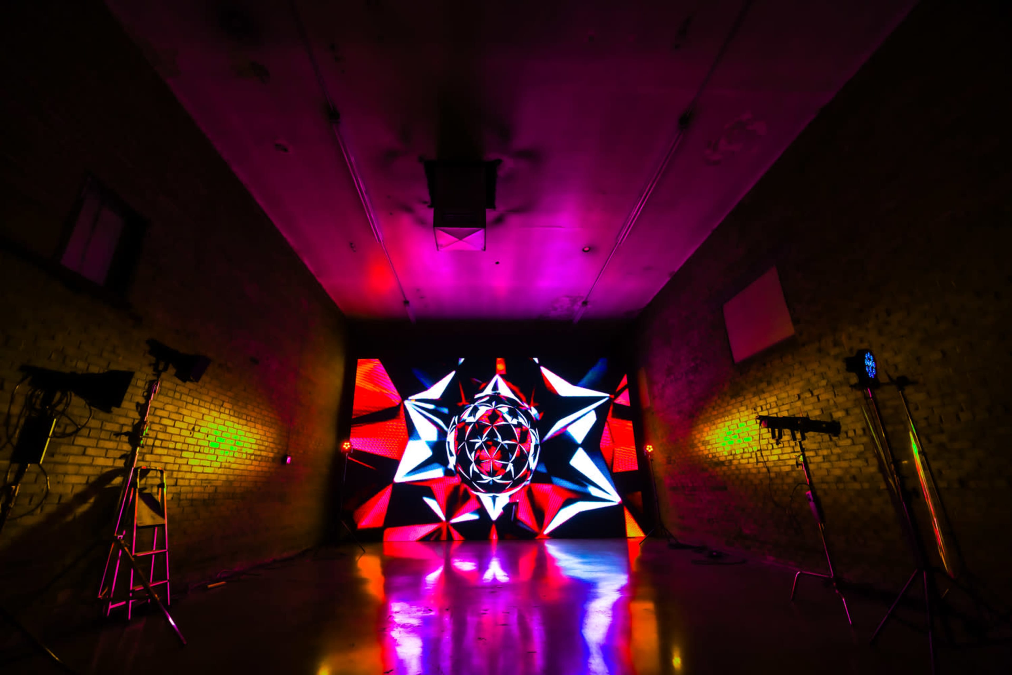 The image shows a dimly lit room with a vibrant, colorful light display featuring geometric patterns on a rear wall and stage lighting equipment set up in the corners.