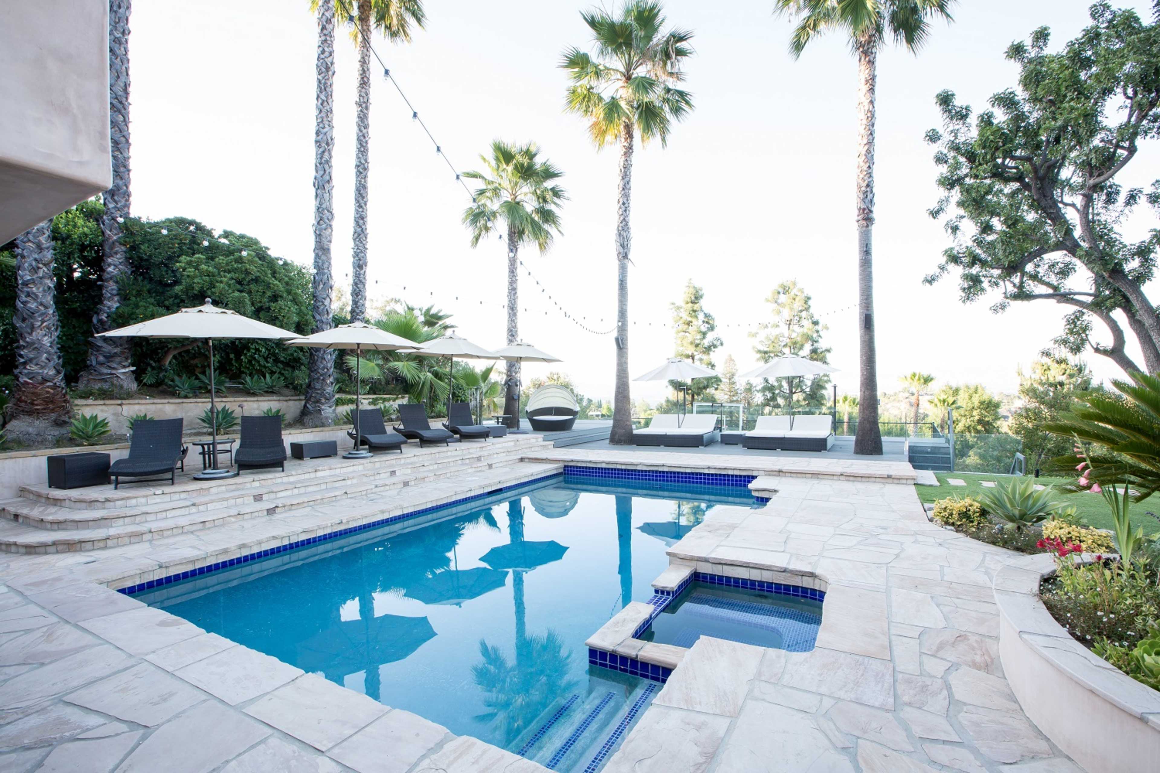 A swimming pool surrounded by palm trees and lounge chairs, with patio umbrellas and a stone deck in a landscaped outdoor area.