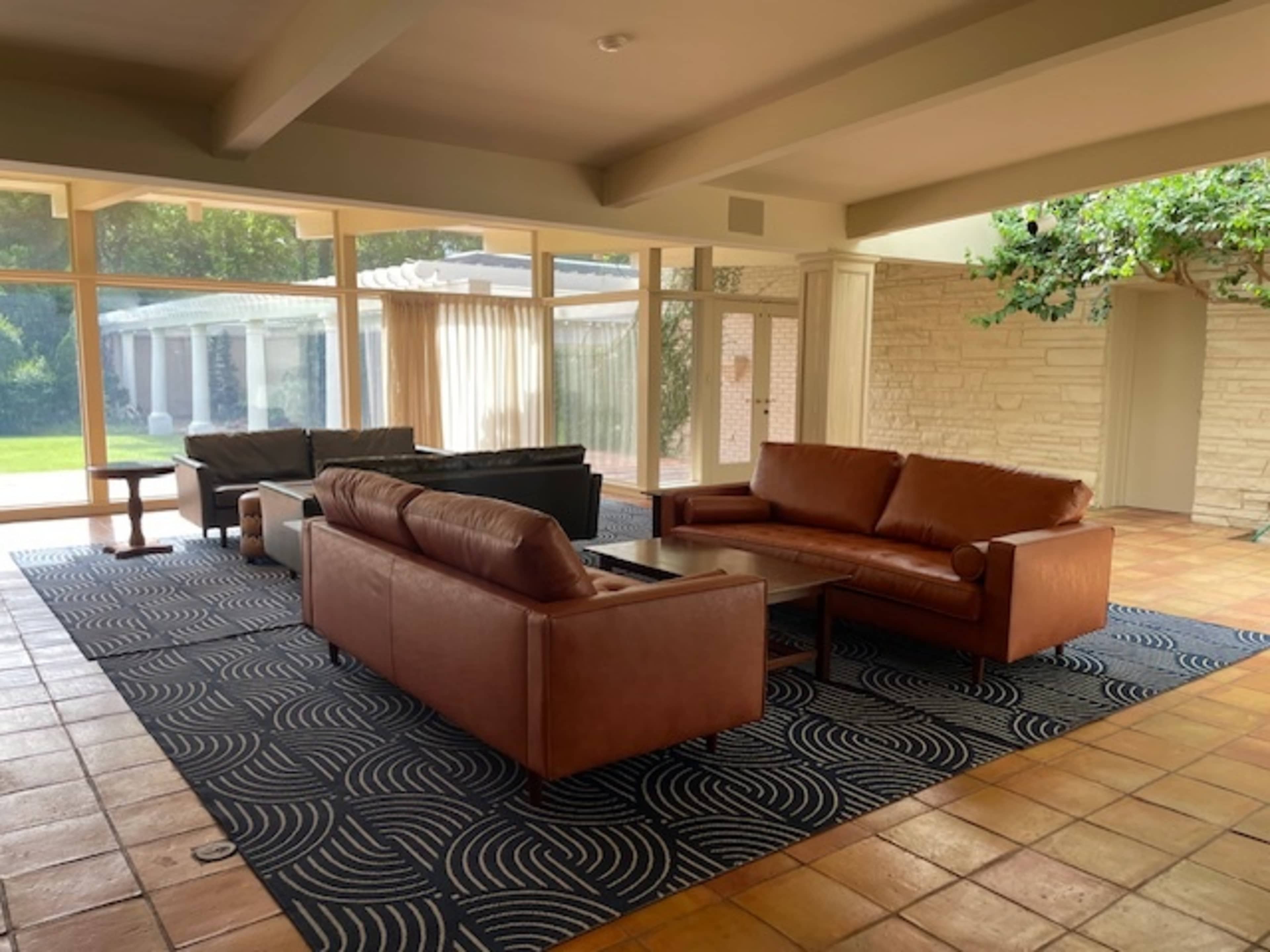 A spacious living room featuring two brown leather sofas and a coffee table on a patterned rug, with large windows allowing natural light to enter.