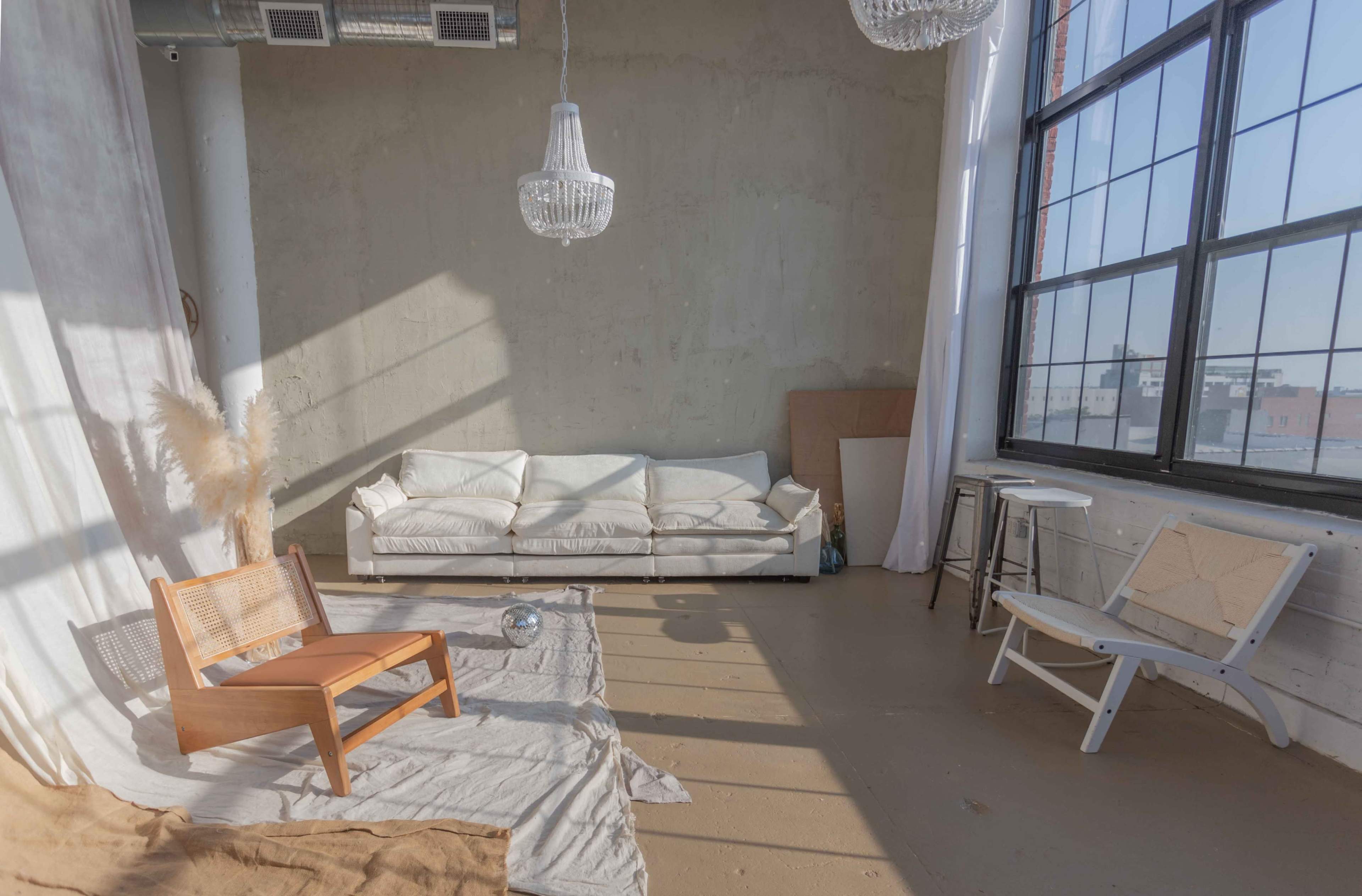 A sunlit room with a white couch, a wooden chair, and large windows, featuring minimal furnishings and light-colored walls.