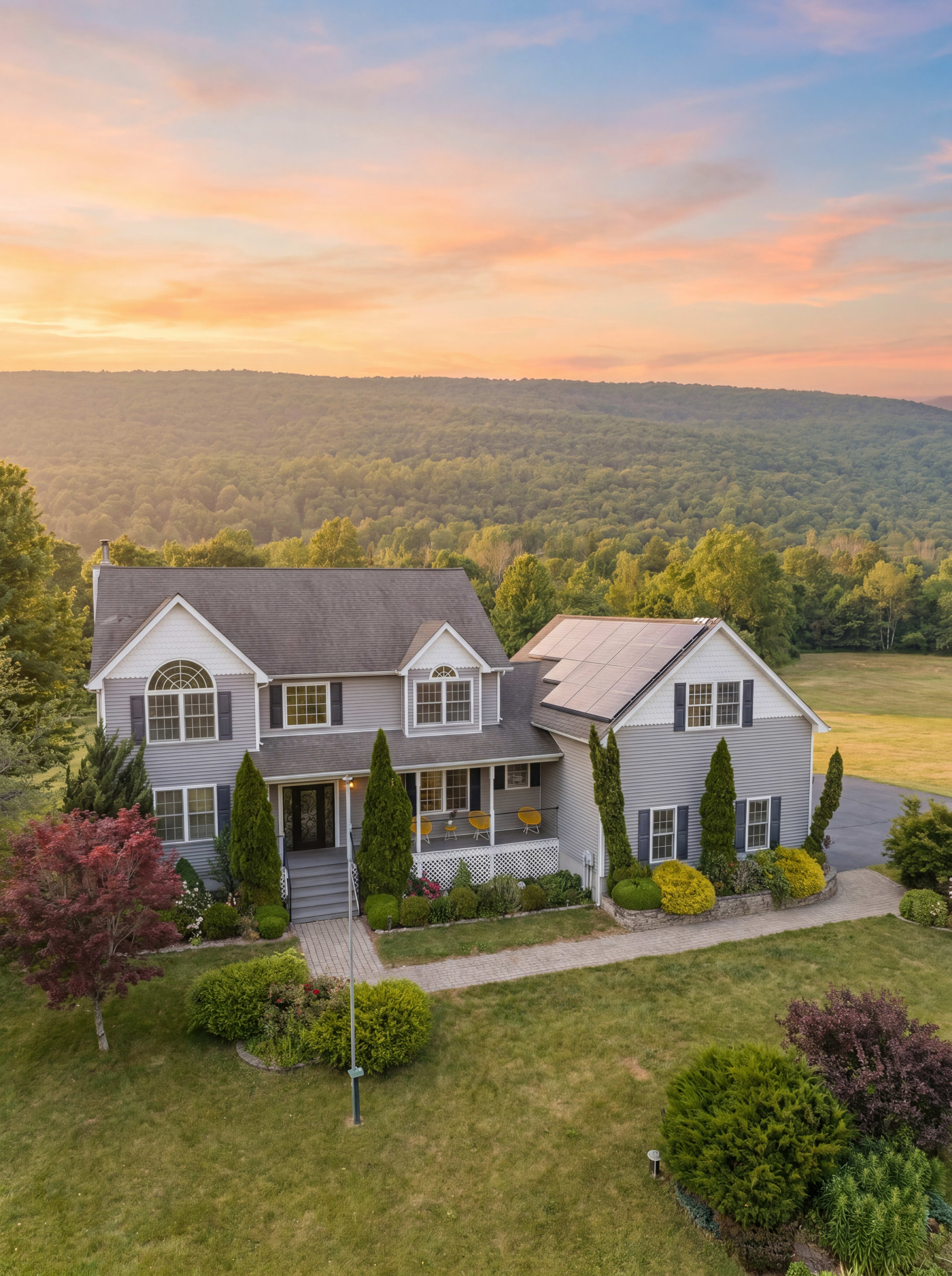 A large gray two-story house with solar panels sits on a well-manicured lawn surrounded by trees and open fields under a colorful sunset.