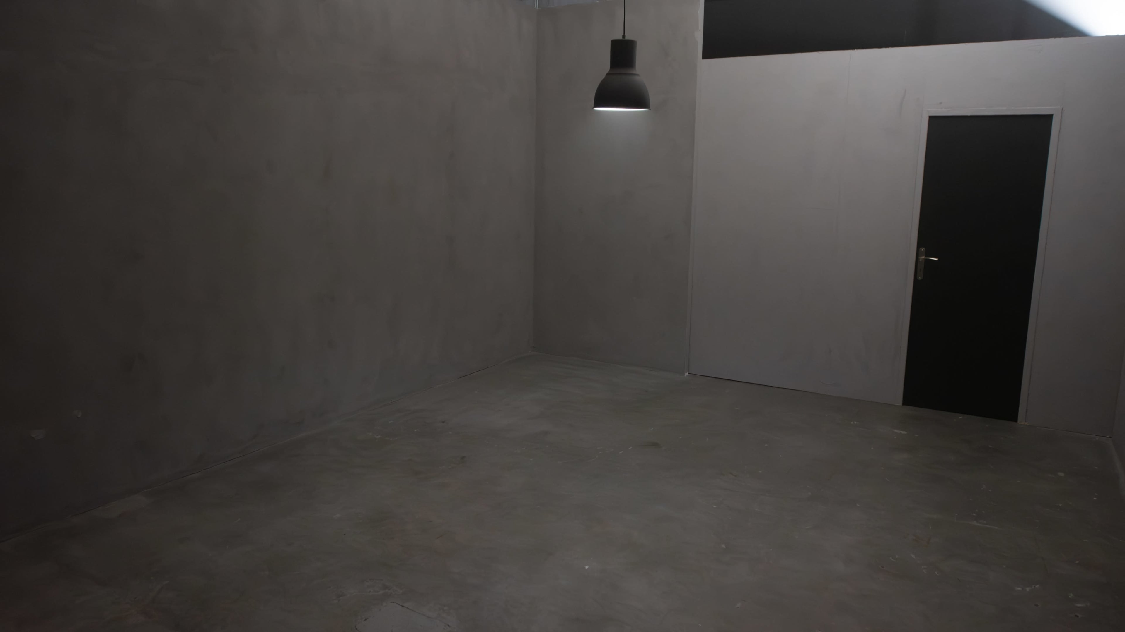 A sparsely furnished, empty room with gray concrete walls and floor, featuring a single light fixture and a black door.