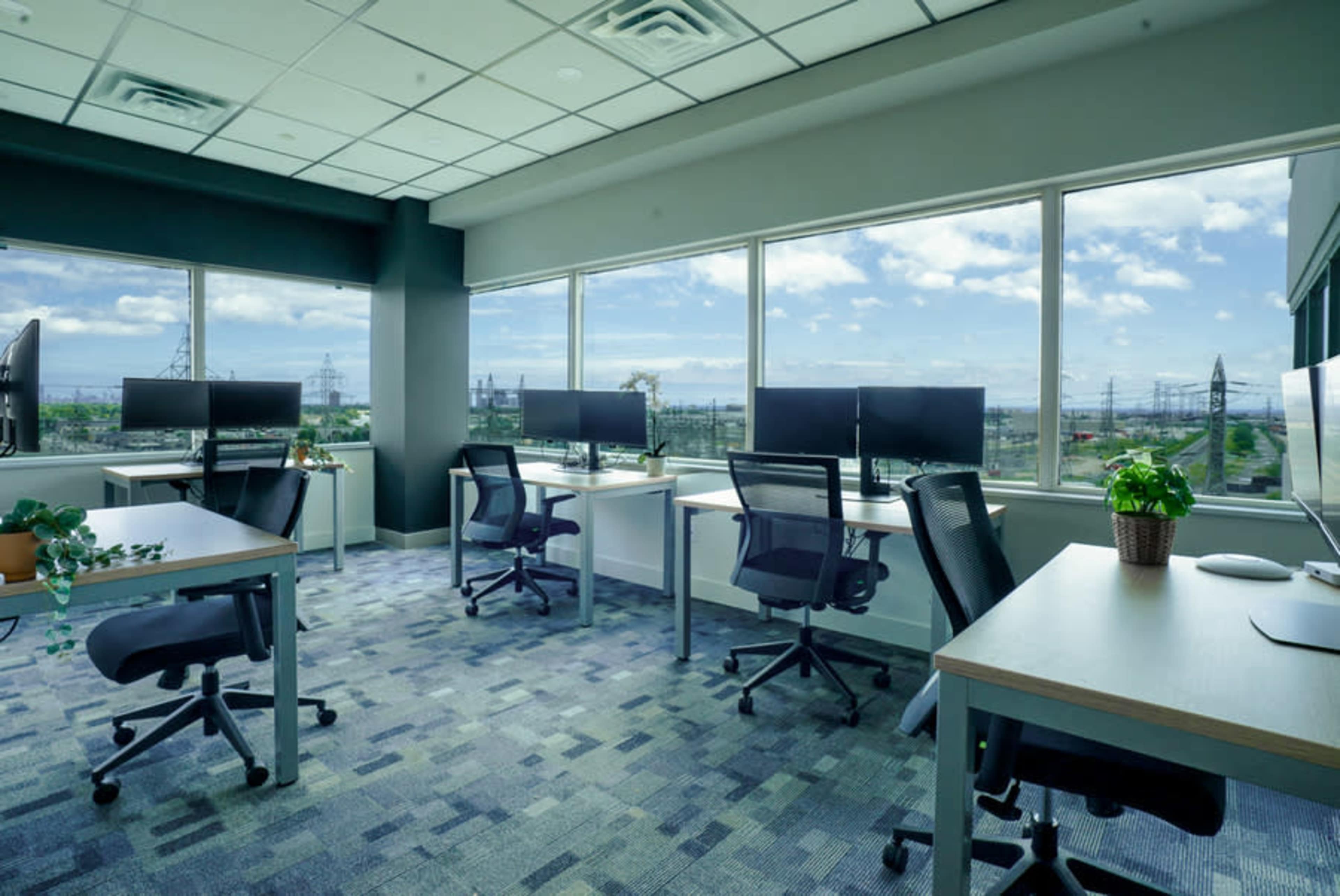 The image shows a modern office space featuring several desks with chairs, multiple computer monitors, and large windows offering a view of the outside.