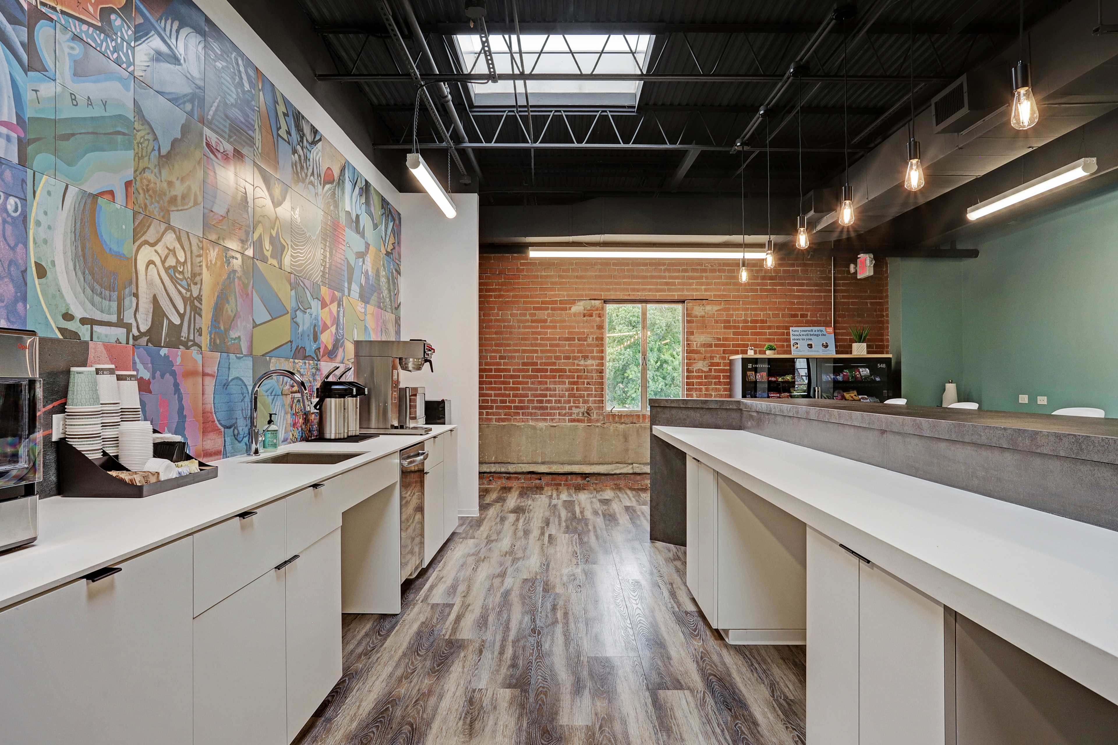A modern kitchen space with a large mural on one wall, coffee machines, and countertops made of various materials, illuminated by pendant lights overhead.