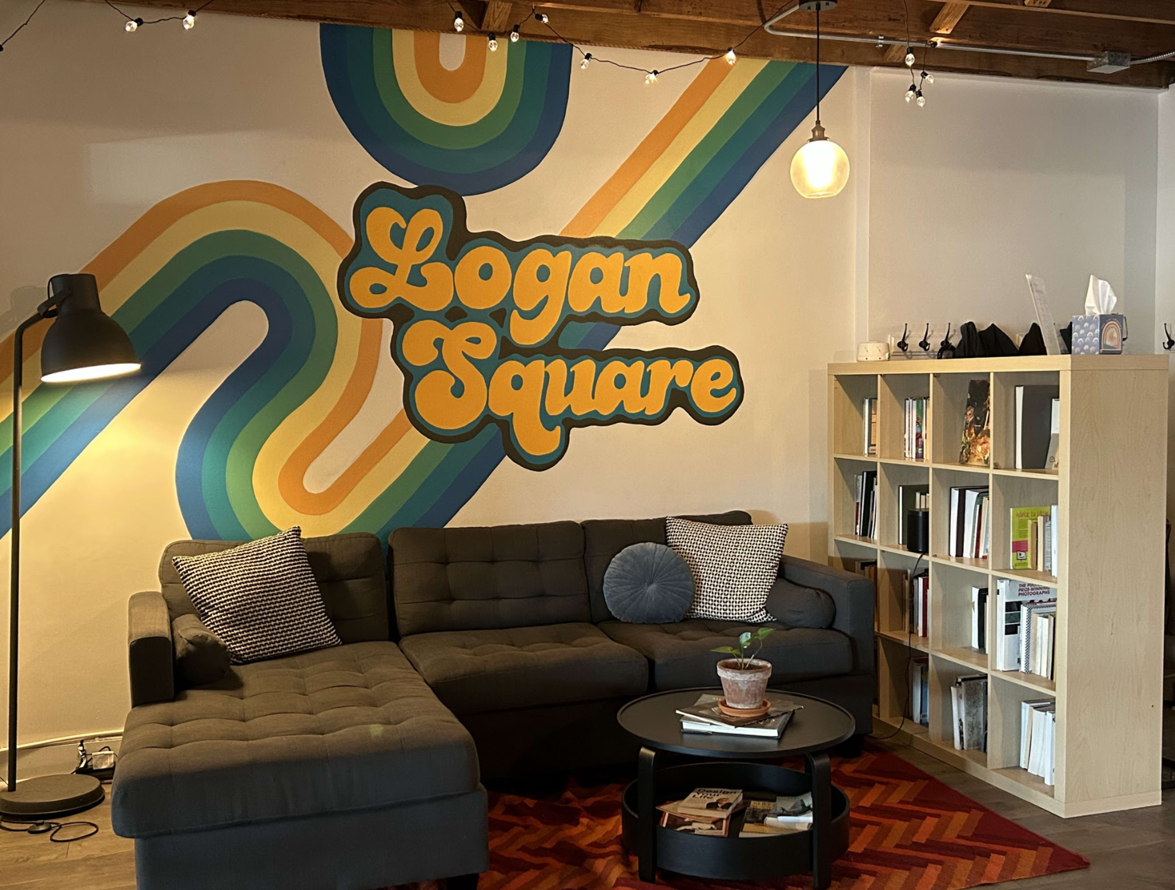 A cozy lounge area featuring a sectional sofa, a coffee table with a plant, and a bookshelf, all against a colorful mural that reads "Logan Square."