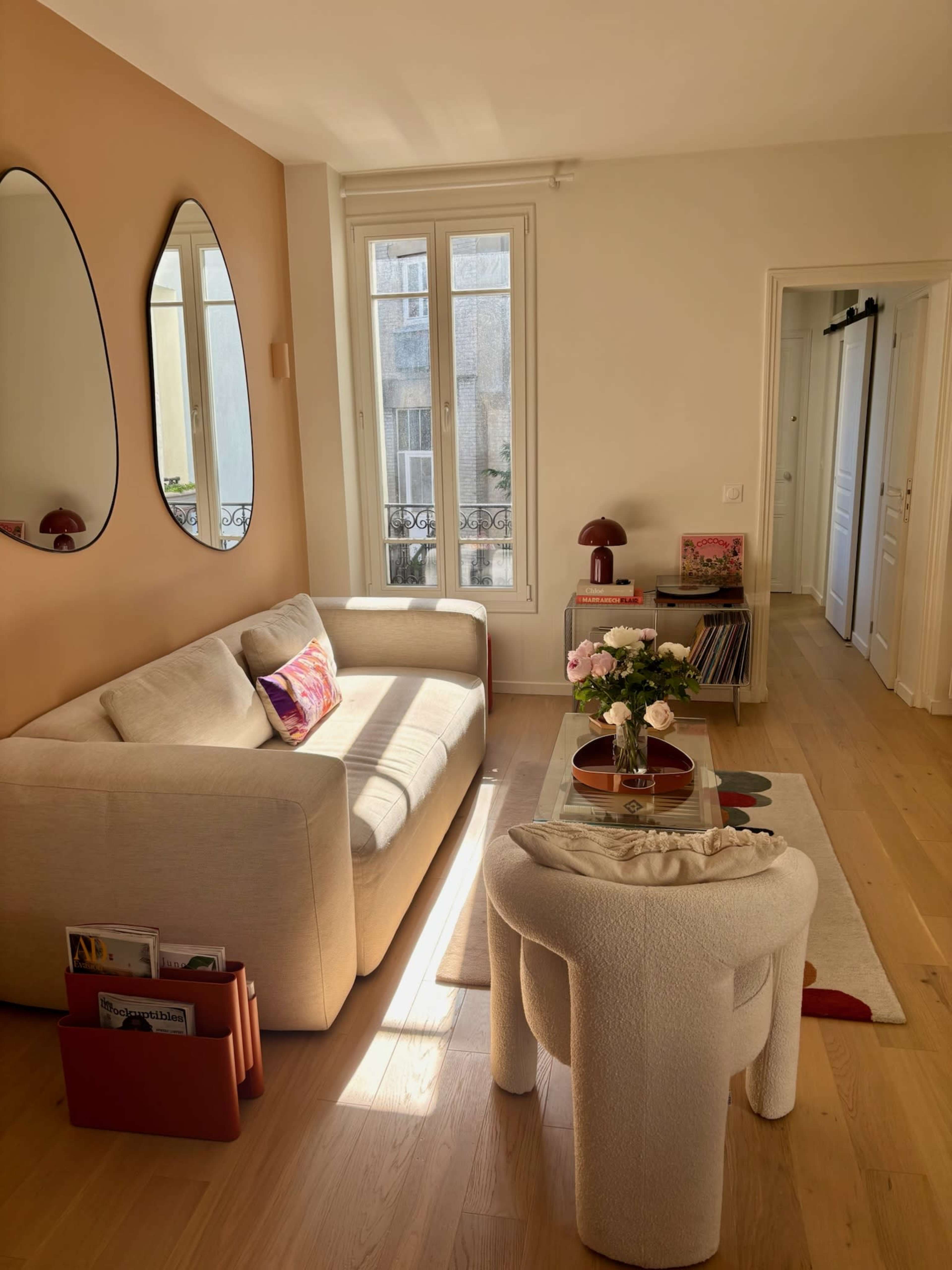The image shows a cozy living room with a beige sofa, a round coffee table, and large mirrors on the wall.