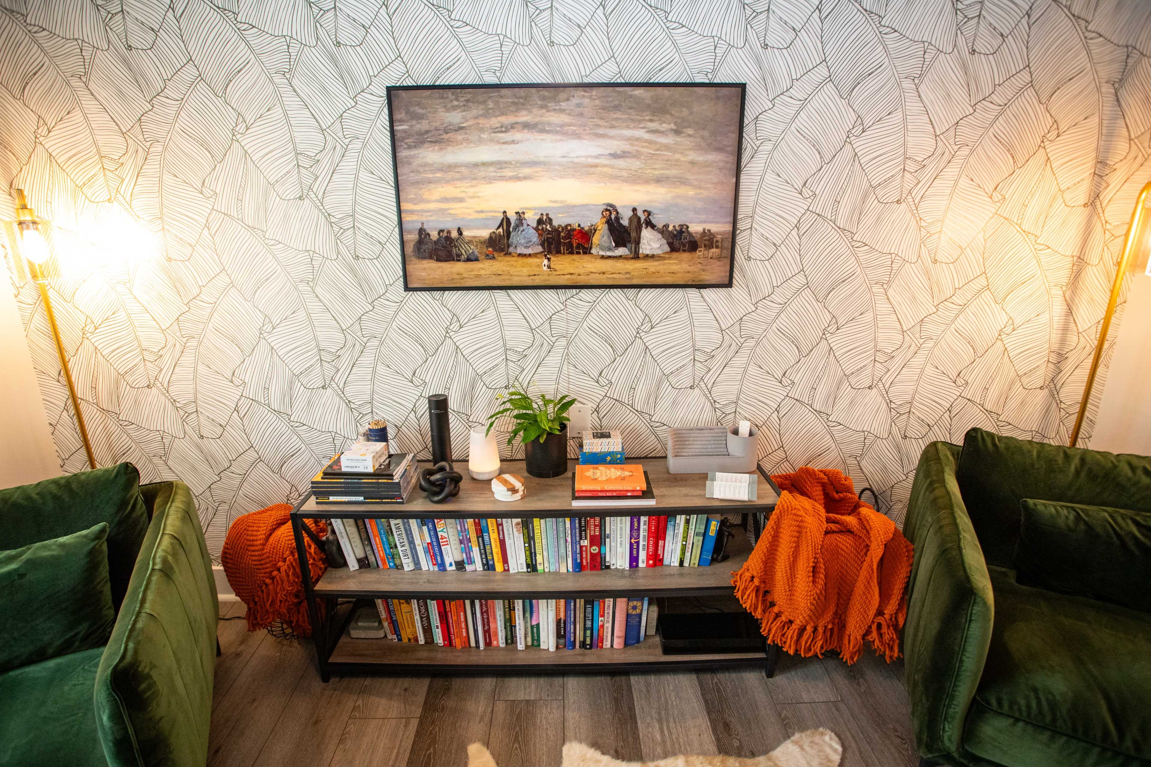 The image shows a stylish living room with two green chairs, a bookshelf filled with books, a table with decorative items, and a large painting on the wall.