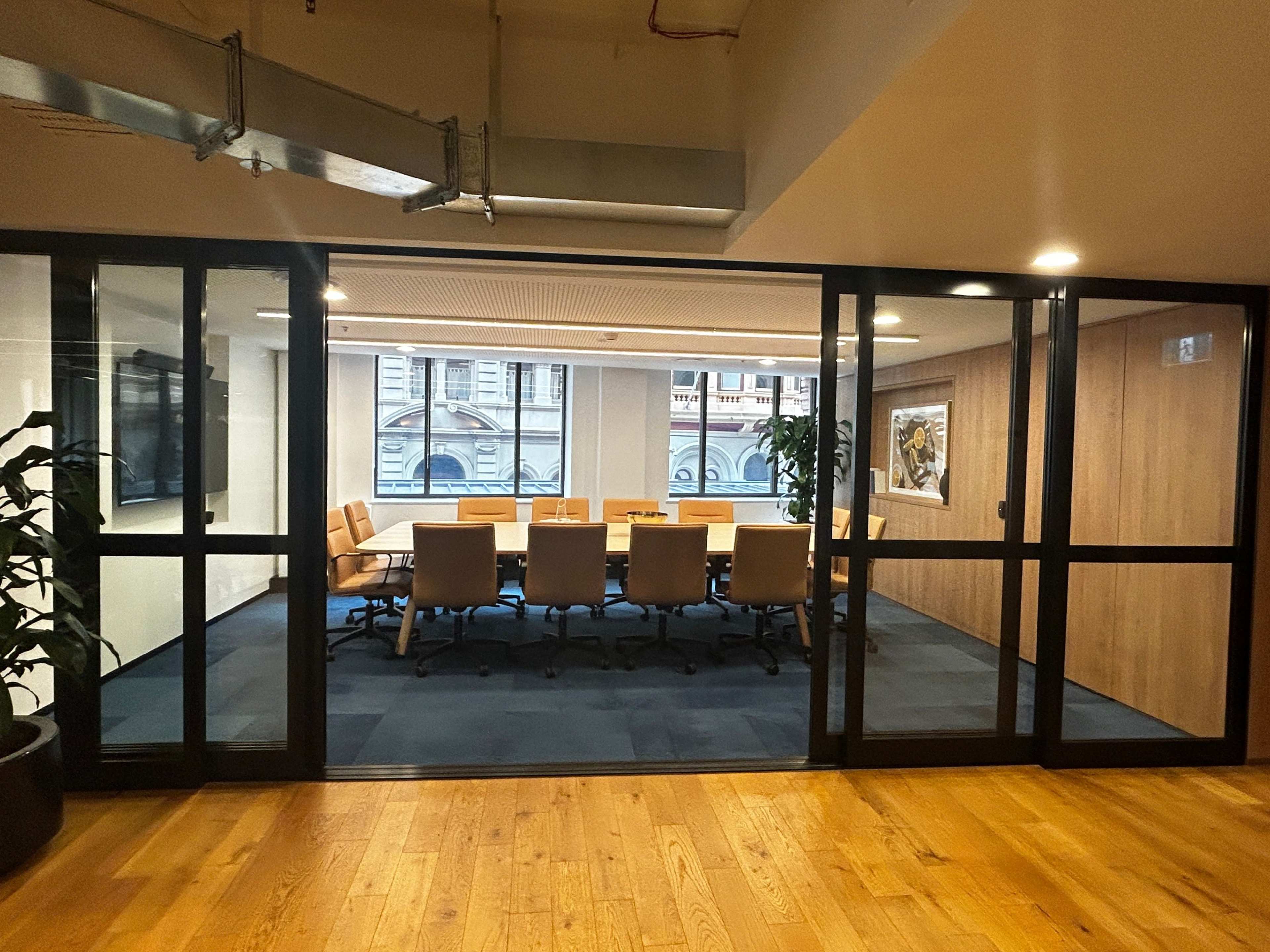 A glass-walled conference room with a long table and several chairs is visible, featuring large windows with a view of a building facade outside.