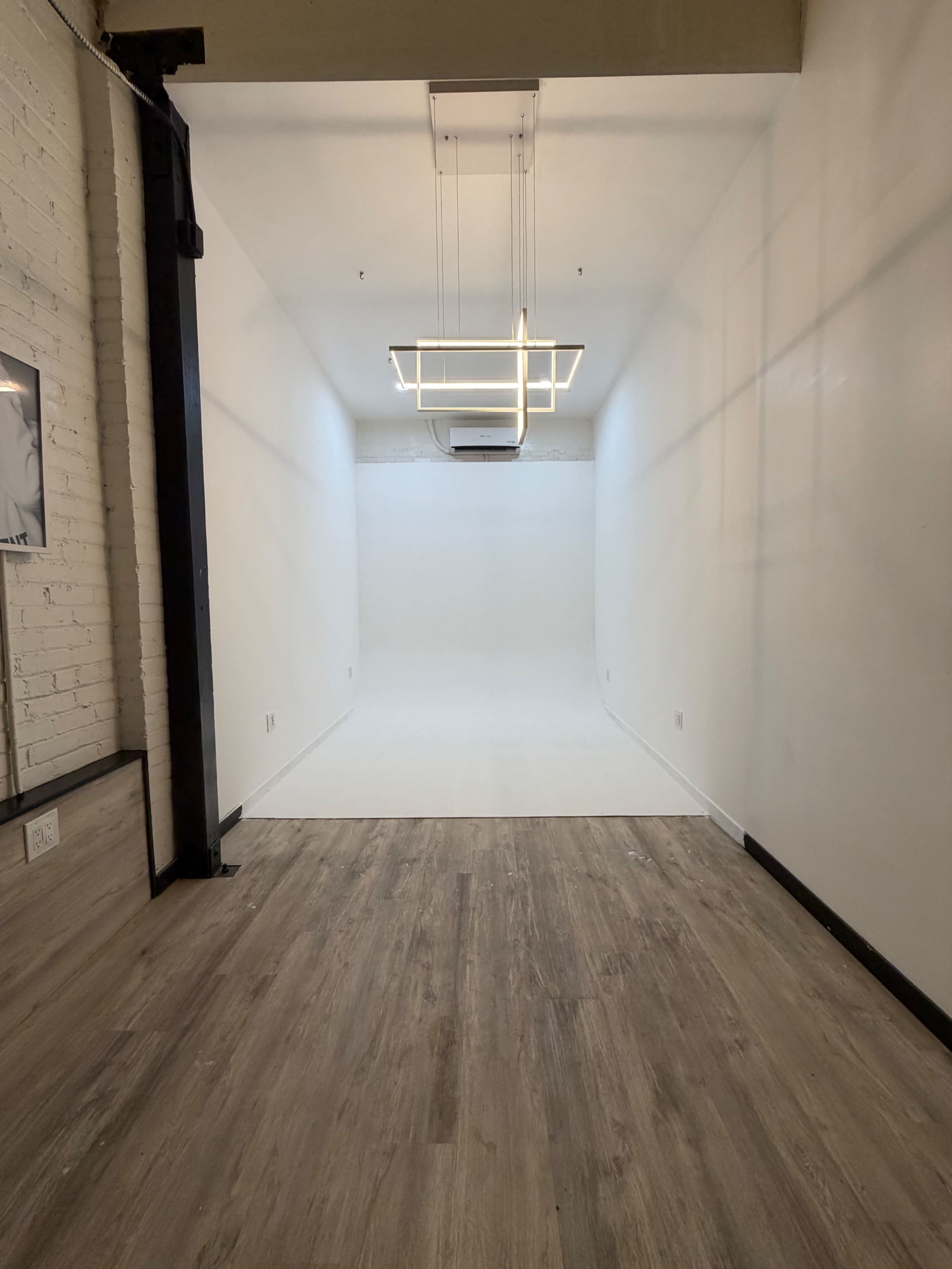The image shows a spacious, empty room with white walls, a wooden floor, and a geometric light fixture hanging from the ceiling.