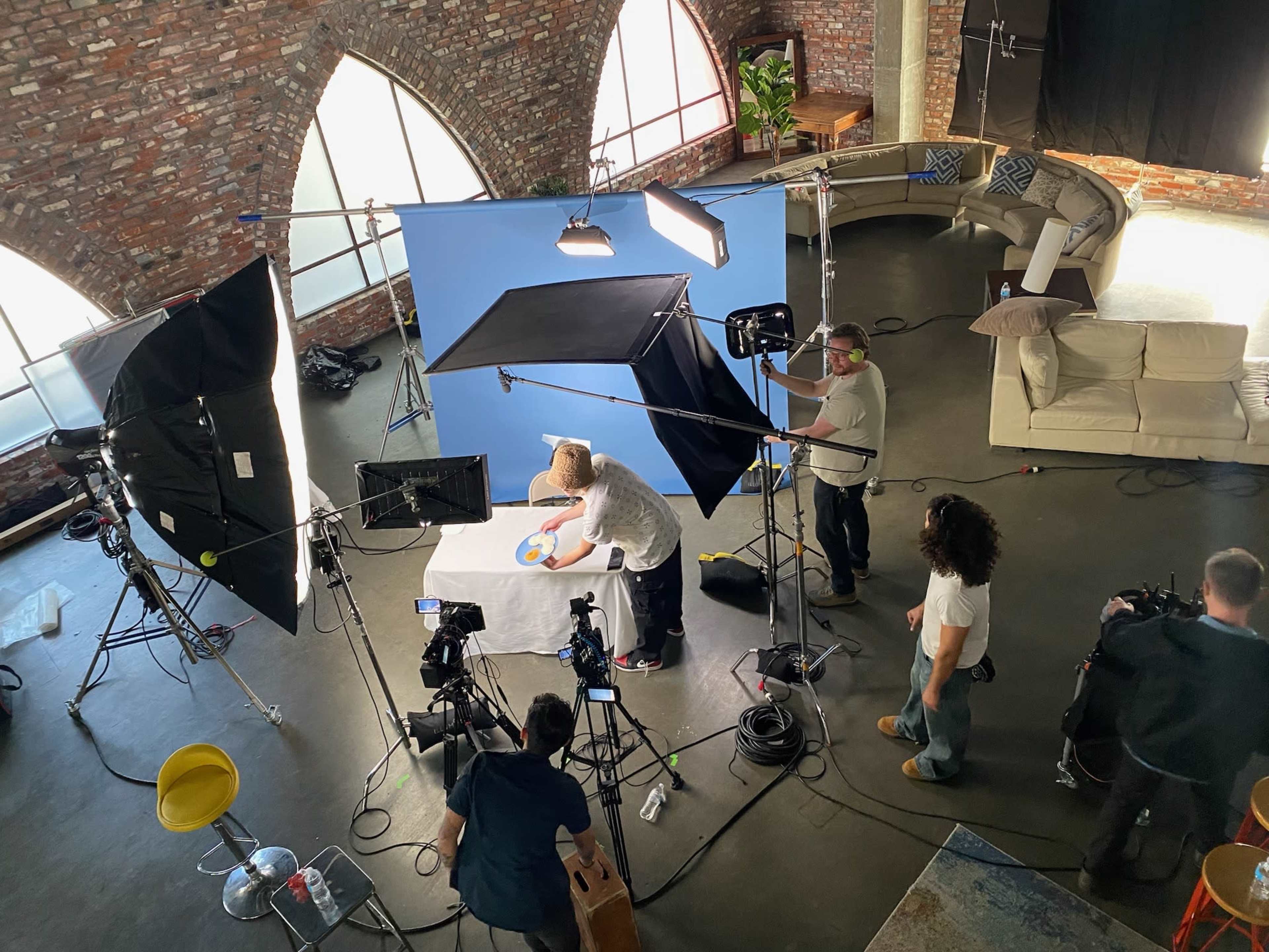 A film crew is set up in a studio with multiple cameras and lights while a person works on a project on a table covered with a white cloth.