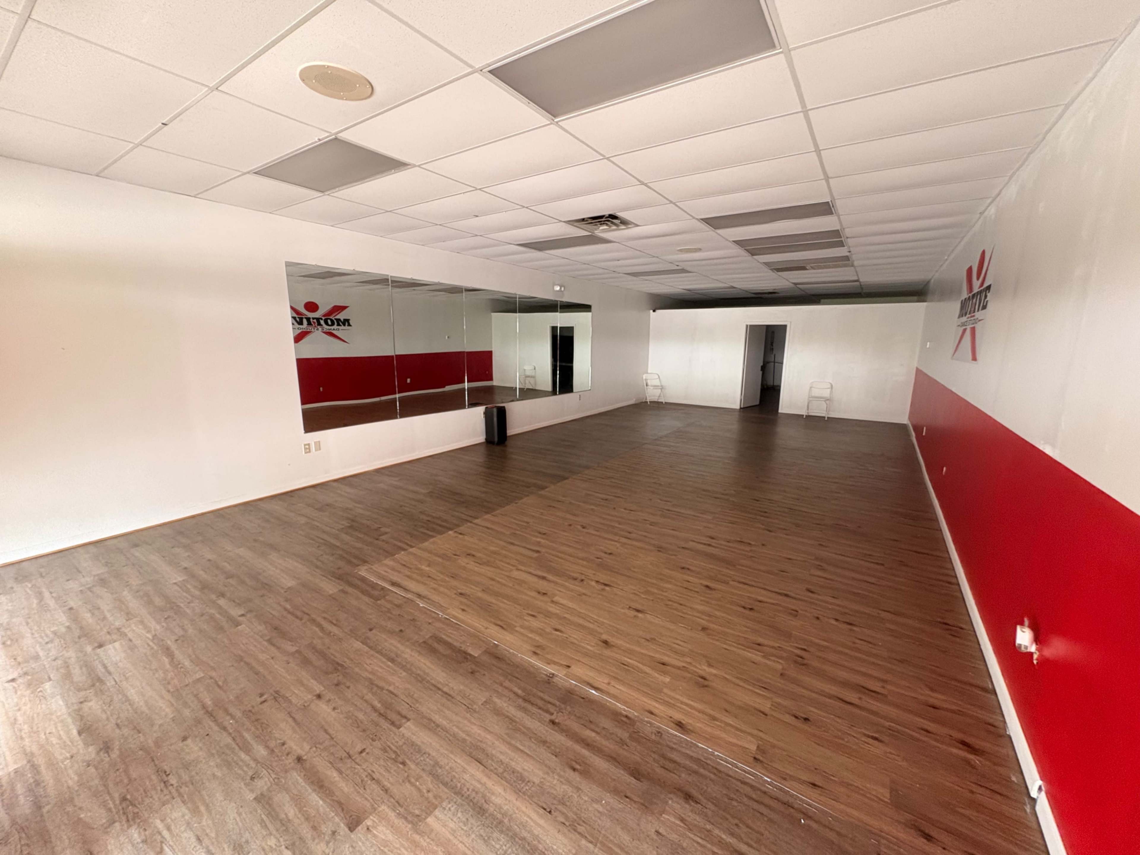 An empty room with wooden flooring features a mirrored wall and red accents, along with two white chairs and a door leading to another space.