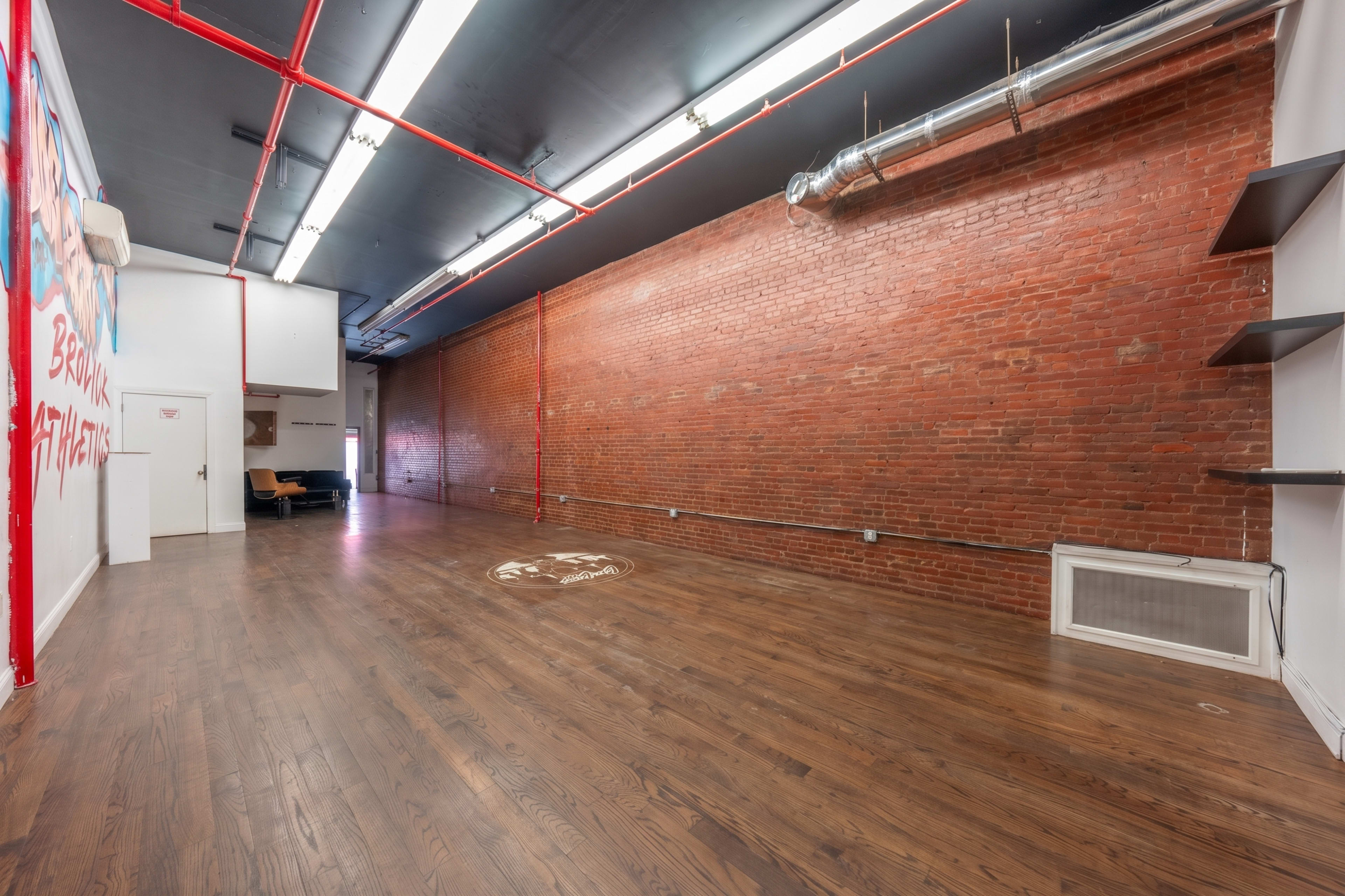 The image shows a spacious, empty room with exposed red brick walls, wooden flooring, and fluorescent lighting.