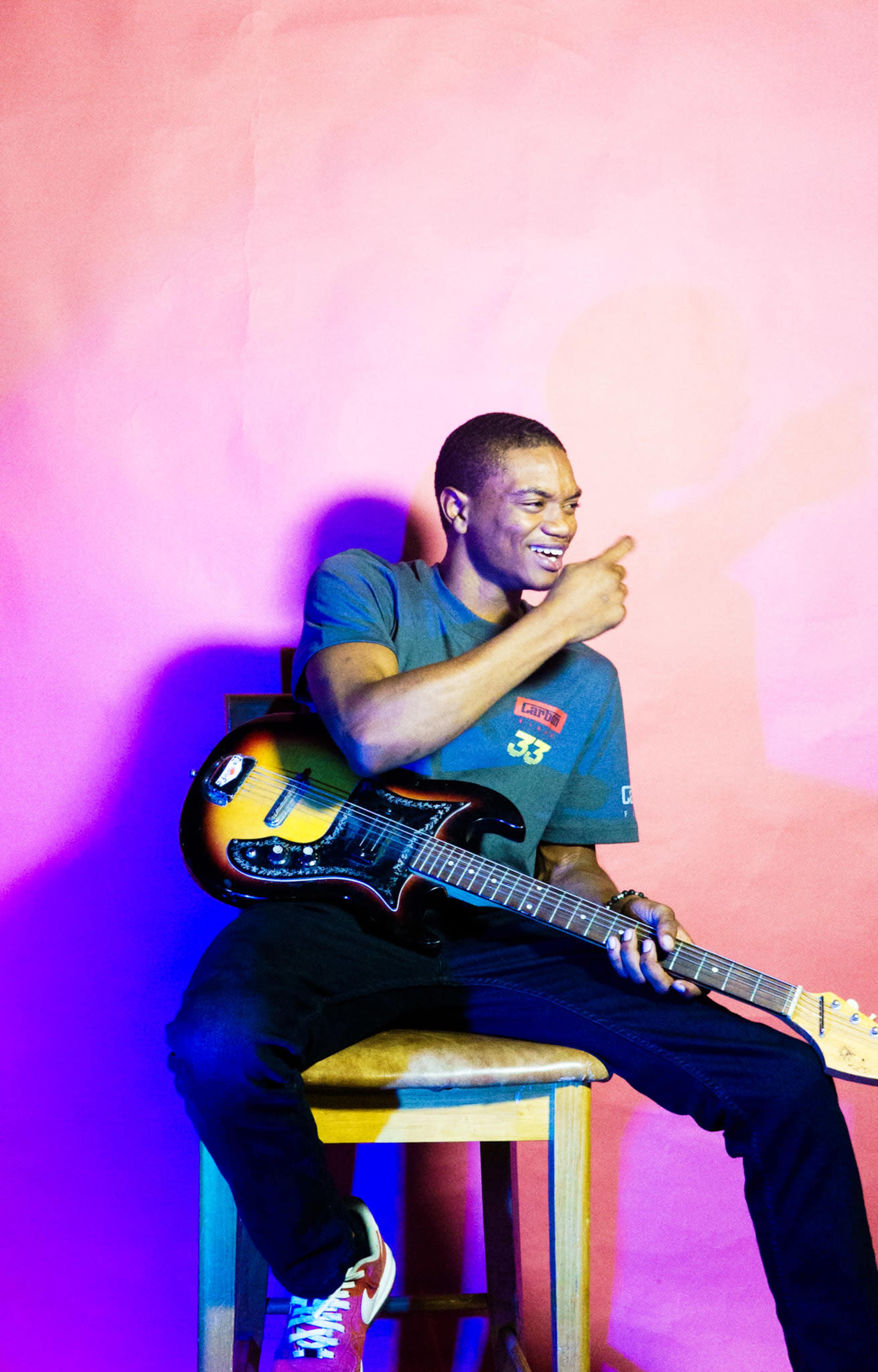 A young man sits on a wooden stool, holding an electric guitar, against a vibrant pink backdrop.