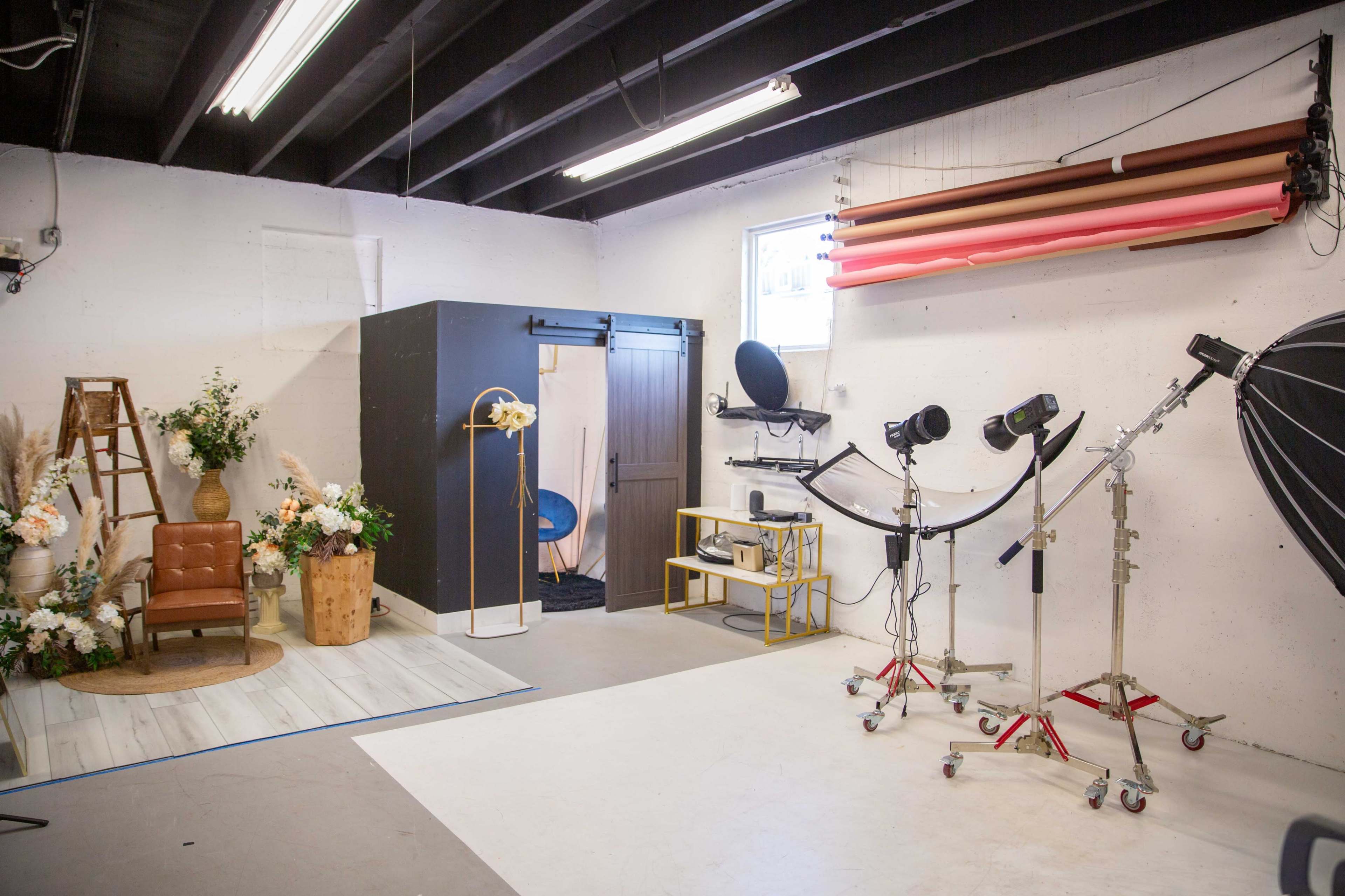 The image shows a photography studio featuring a floral backdrop, a brown chair, lighting equipment, and various props arranged for a shoot.