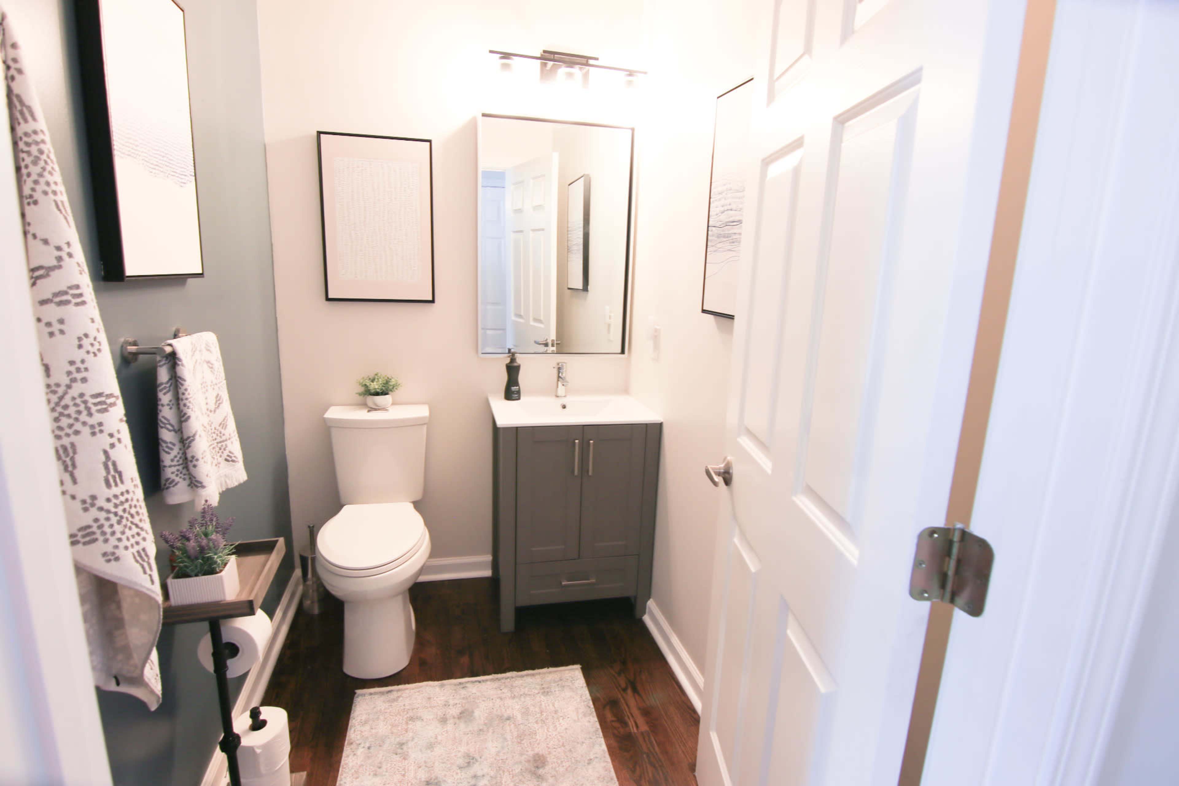 The image shows a compact bathroom featuring a white toilet, a gray vanity with a sink, a wall mirror, and decorative wall art.