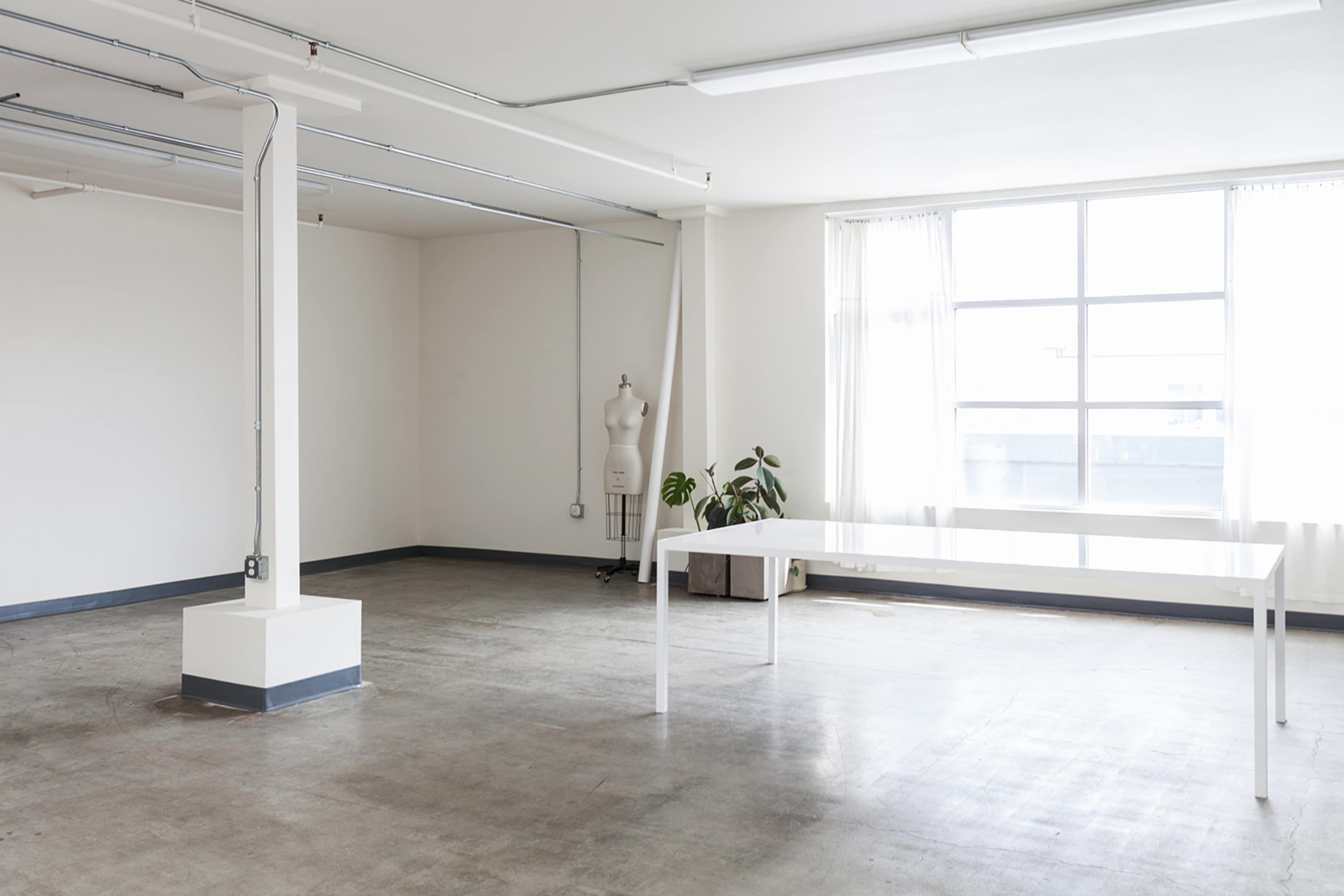 A spacious, empty room with a tall window, a plant, a table, and a sewing mannequin.