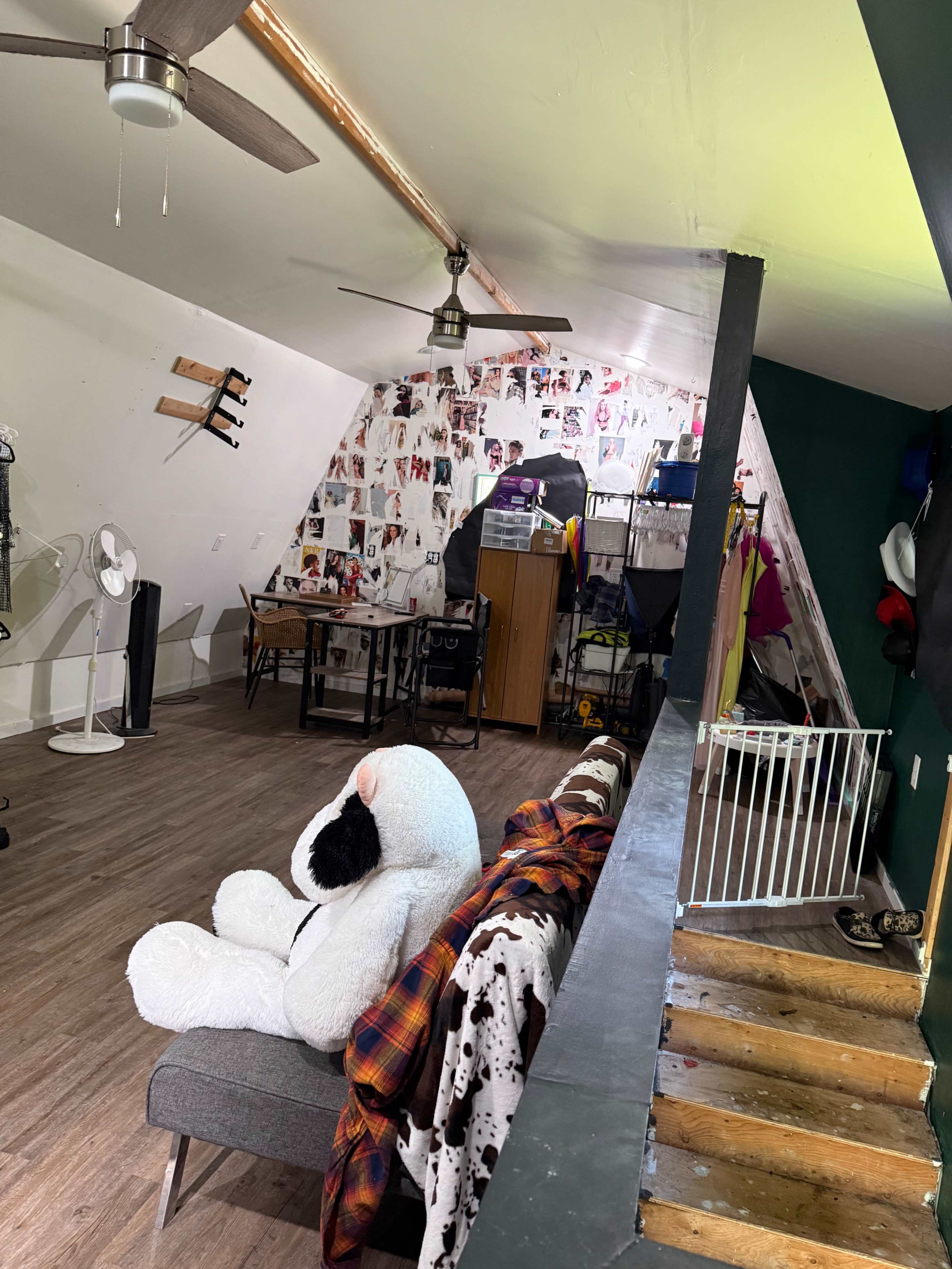 The image shows a room with sloped ceilings, featuring a large stuffed animal on a couch, a table covered with various items, and posters on the walls.