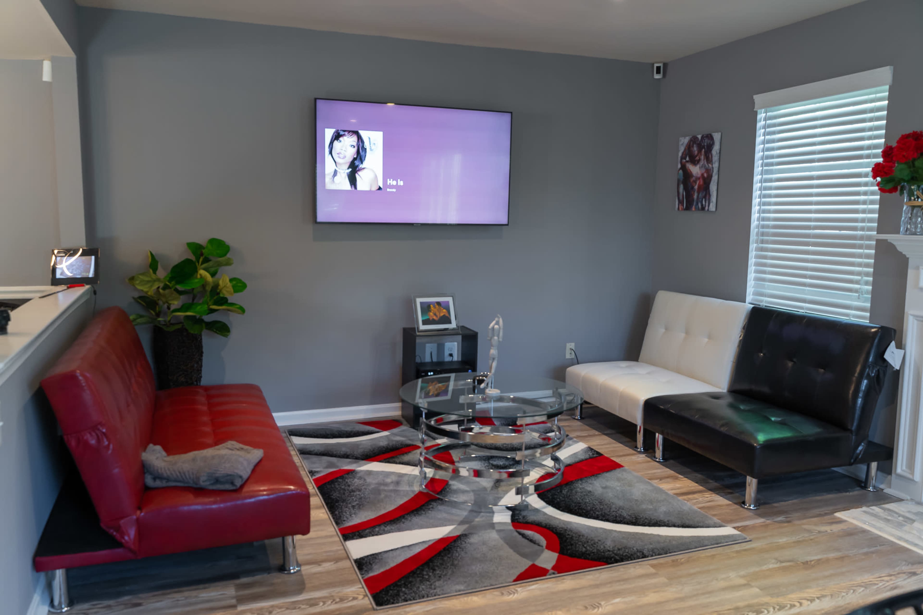 A modern living room featuring a red sofa, a black and white sofa, a glass coffee table on a patterned rug, and a wall-mounted television.
