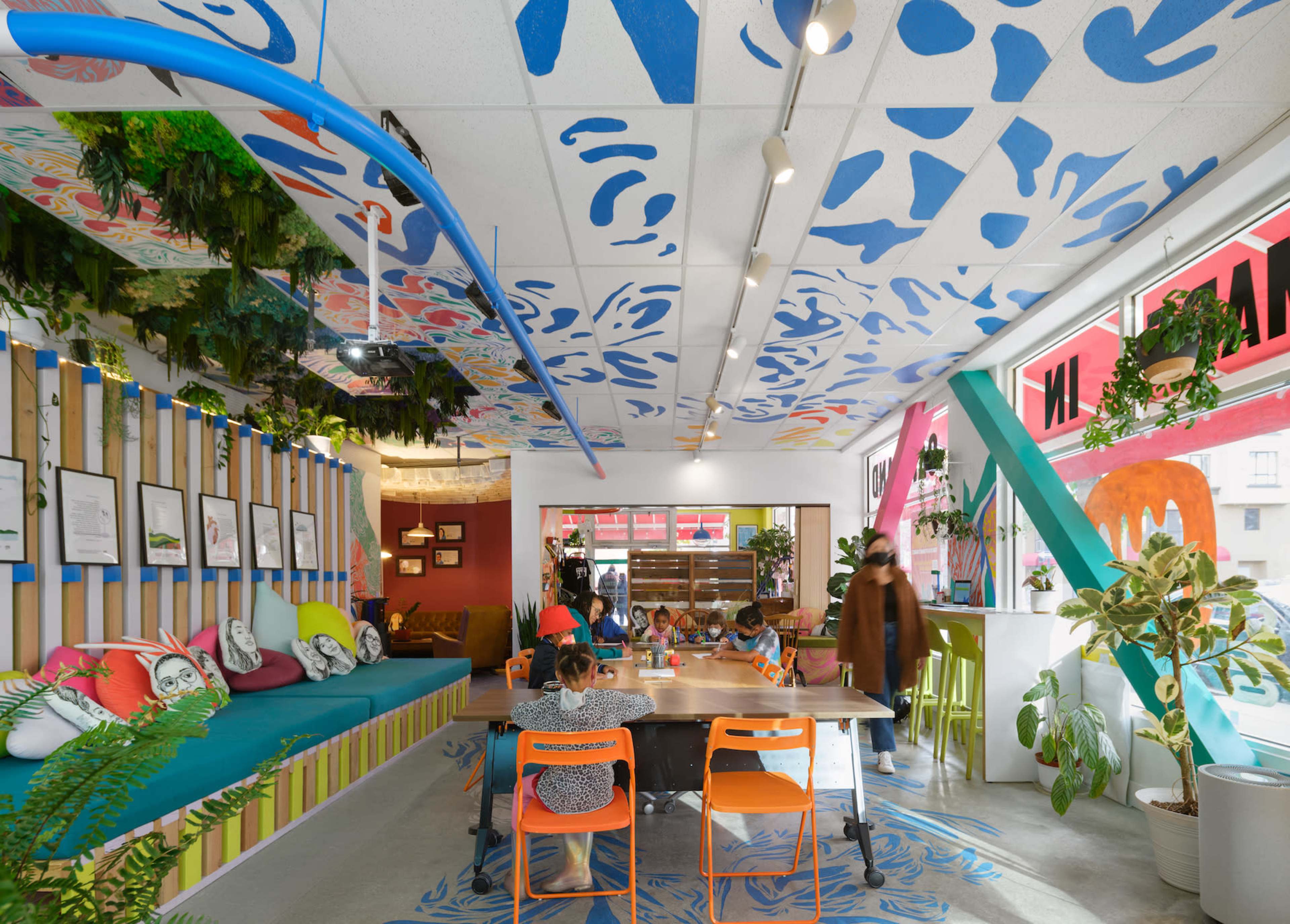 A vibrant café interior featuring colorful wall art, a mix of seating options, and plants, with patrons engaging in conversation at communal tables.