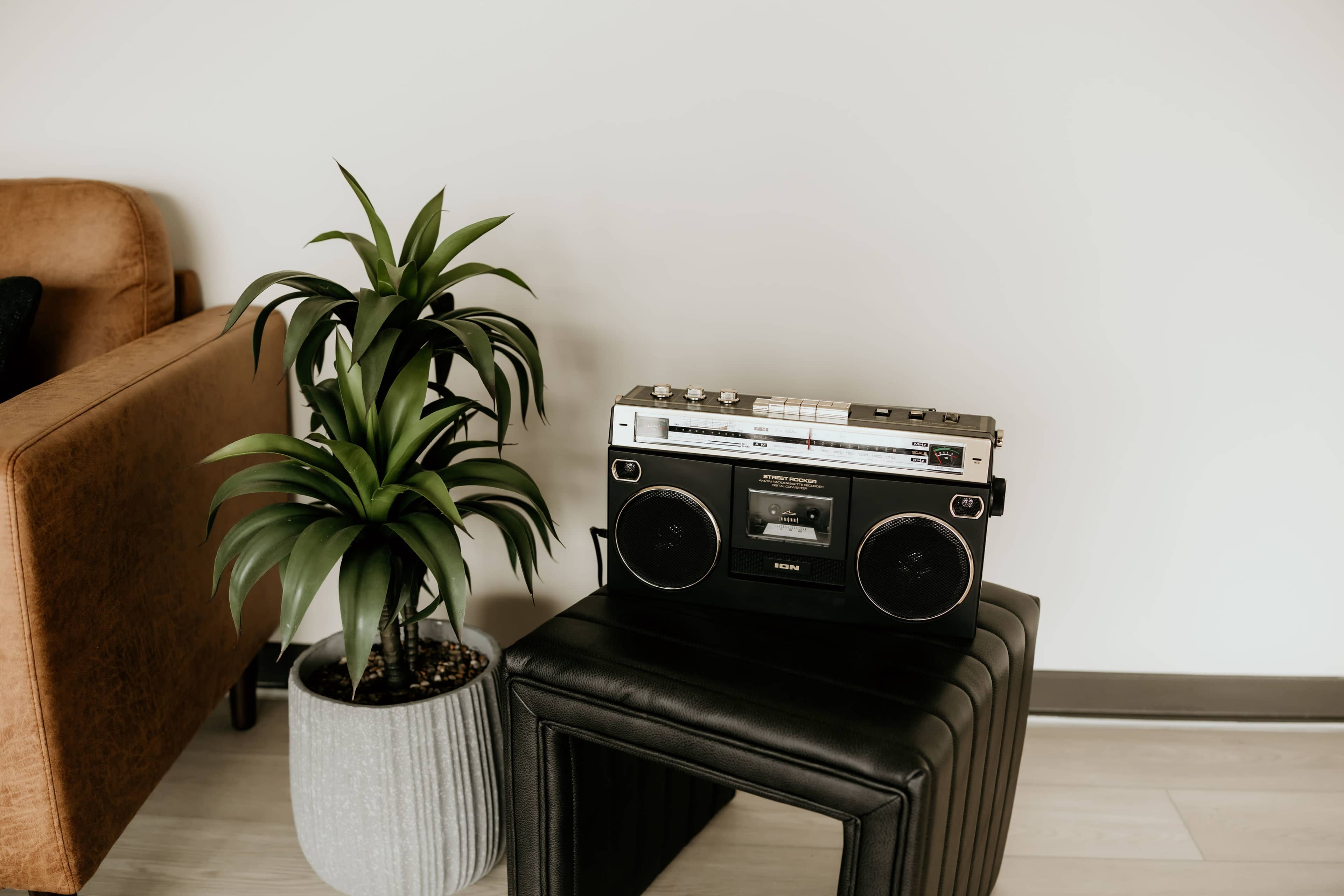 A vintage boombox sits on a black ottoman next to a potted plant and a brown sofa.