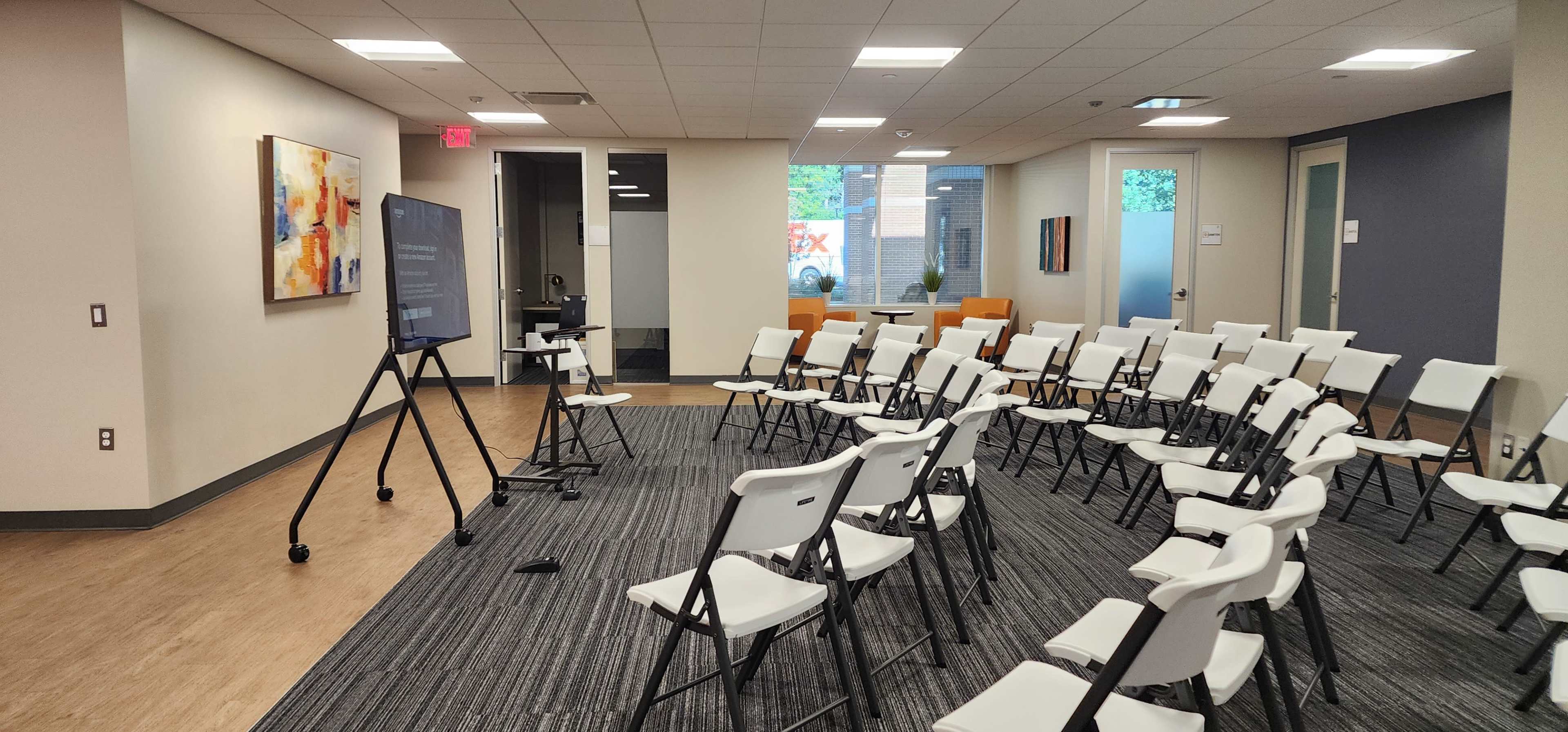 The image shows a conference room set up with white folding chairs arranged in rows facing a large screen on a stand.