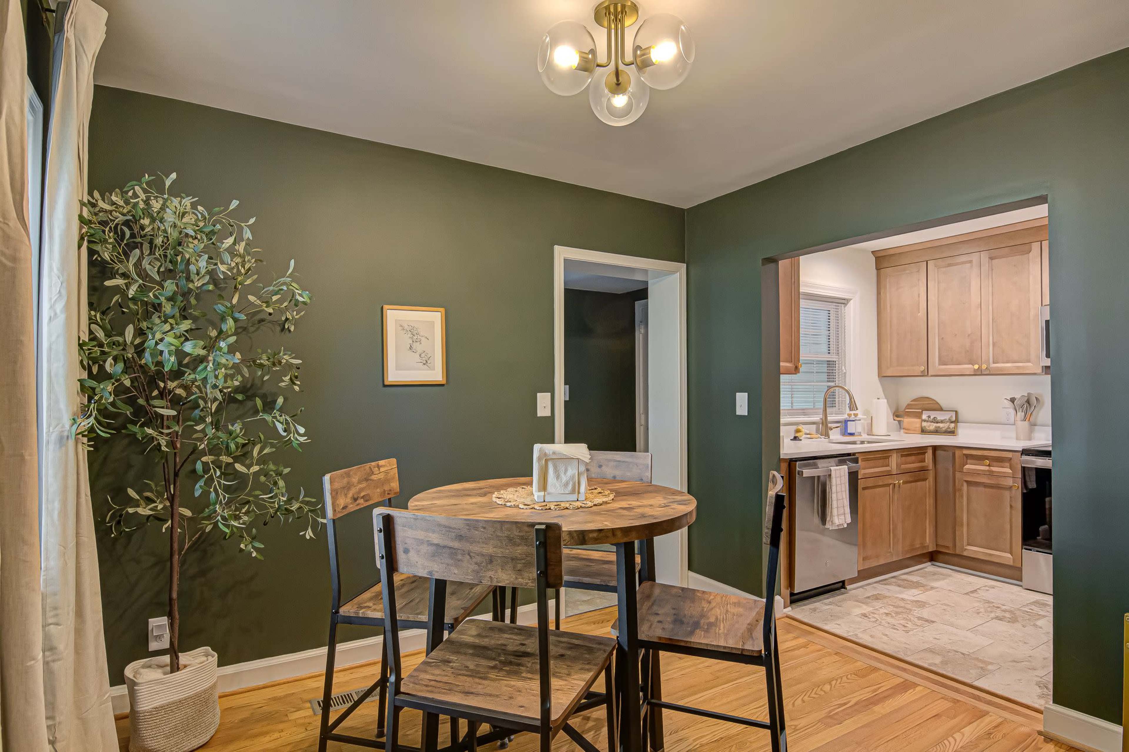 The image shows a dining area featuring a round wooden table with four chairs, a potted plant, and an open kitchen visible through a doorway.
