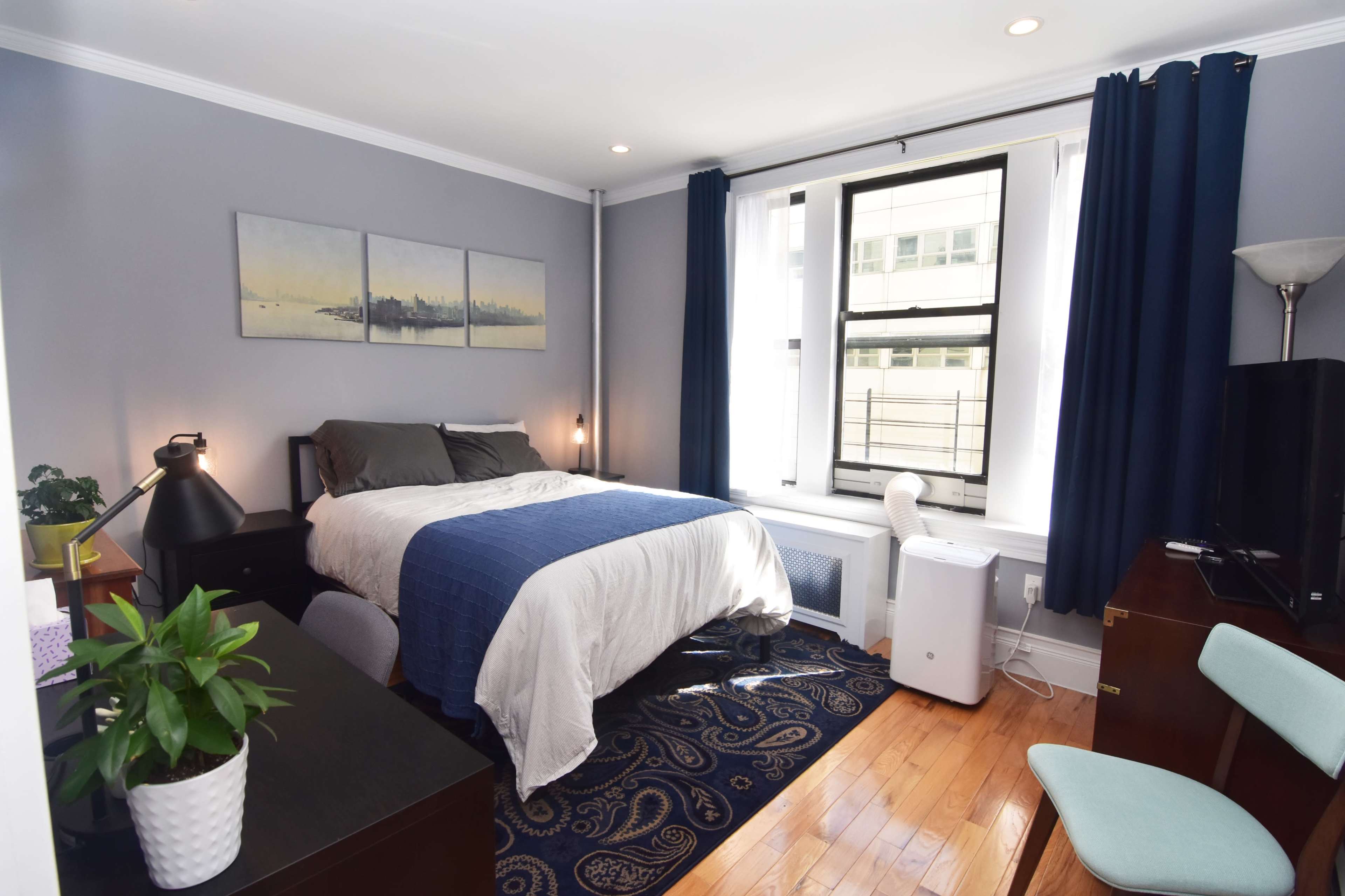 The image shows a neatly arranged bedroom featuring a bed, desk, plant, and window with curtains.