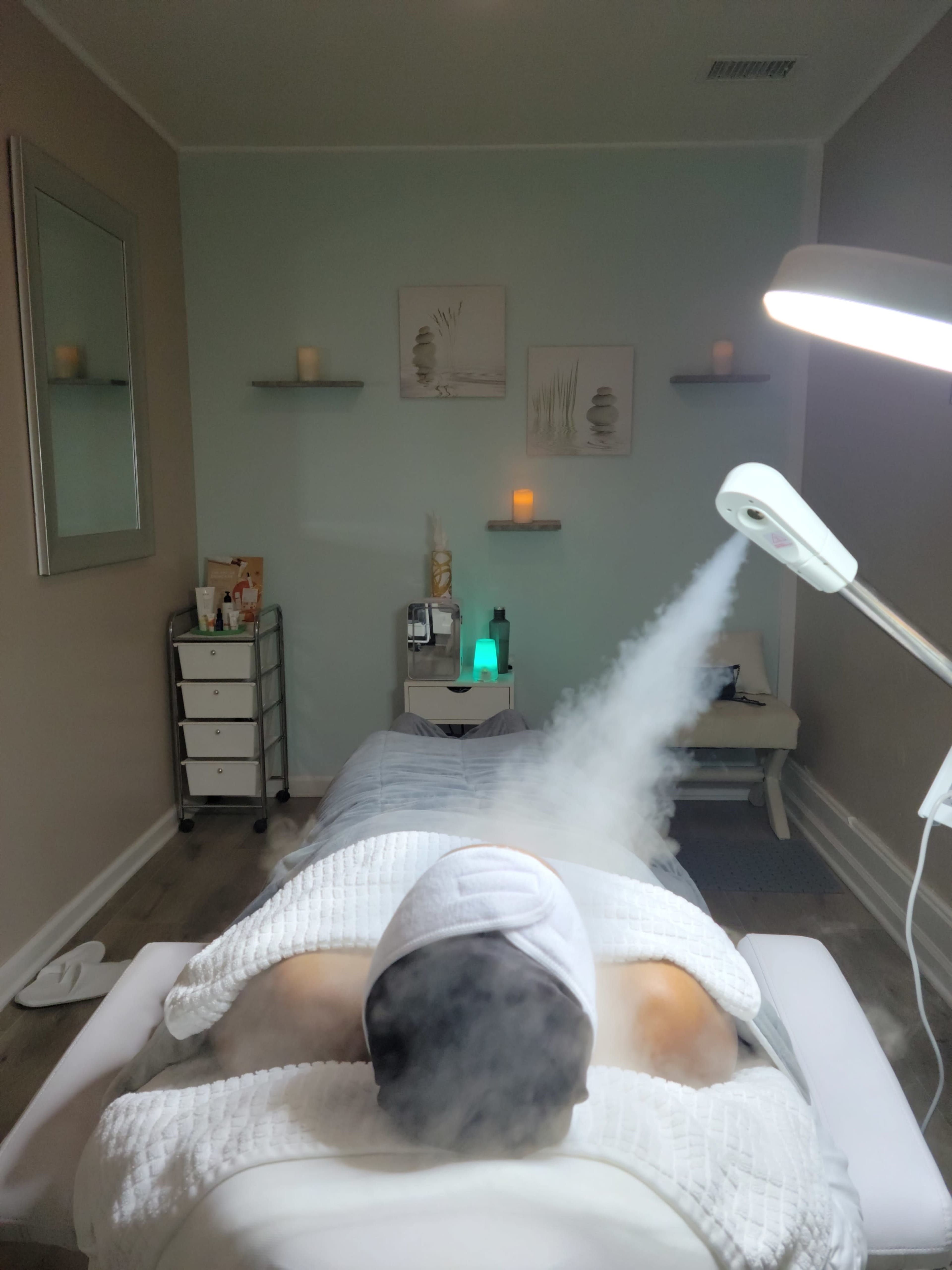 A person lies on a treatment table in a spa room while steam is applied to their face.