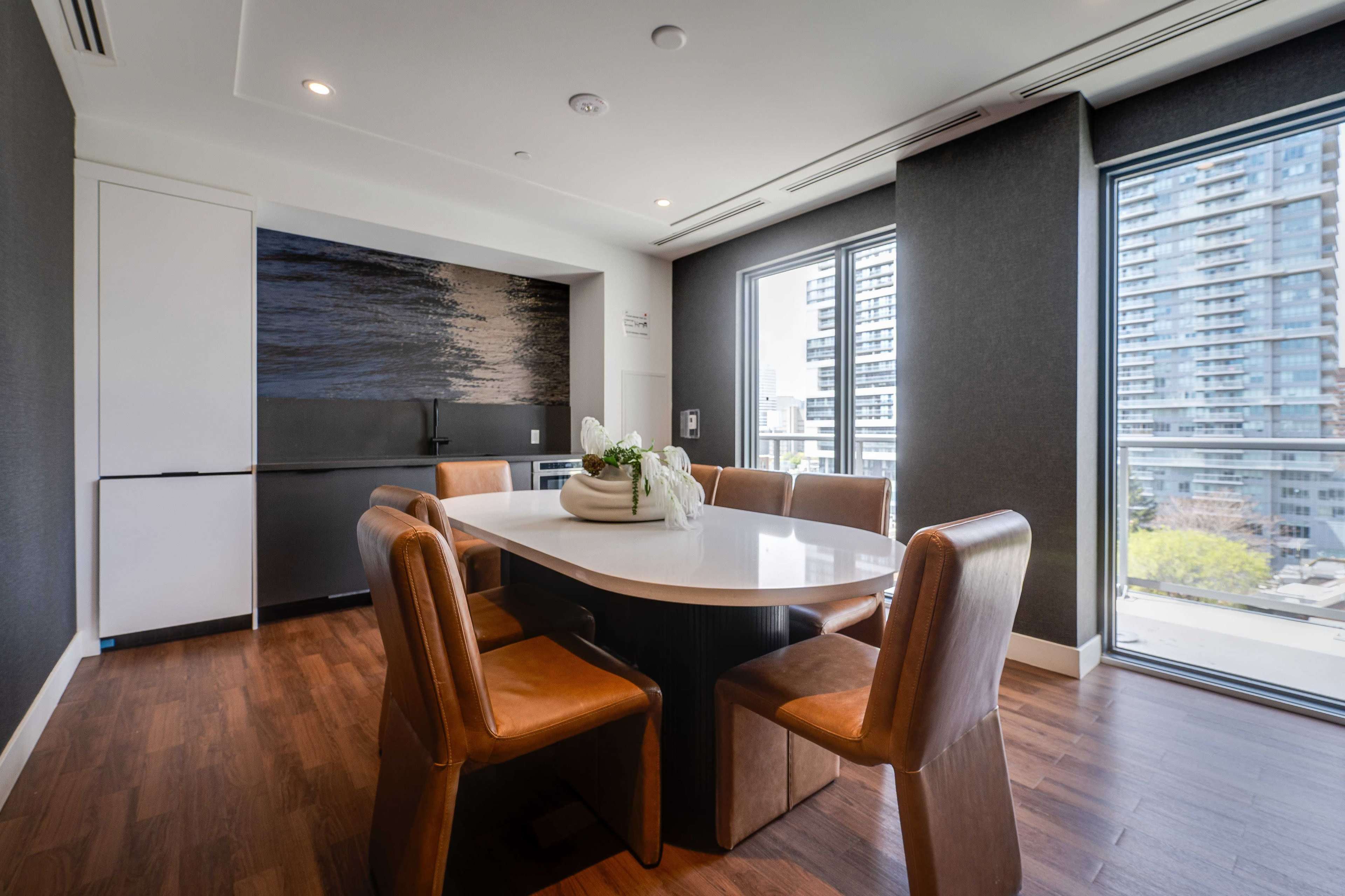 The image shows a modern dining area with a round table surrounded by brown upholstered chairs, large windows offering a view of a cityscape, and a textured wall art feature.
