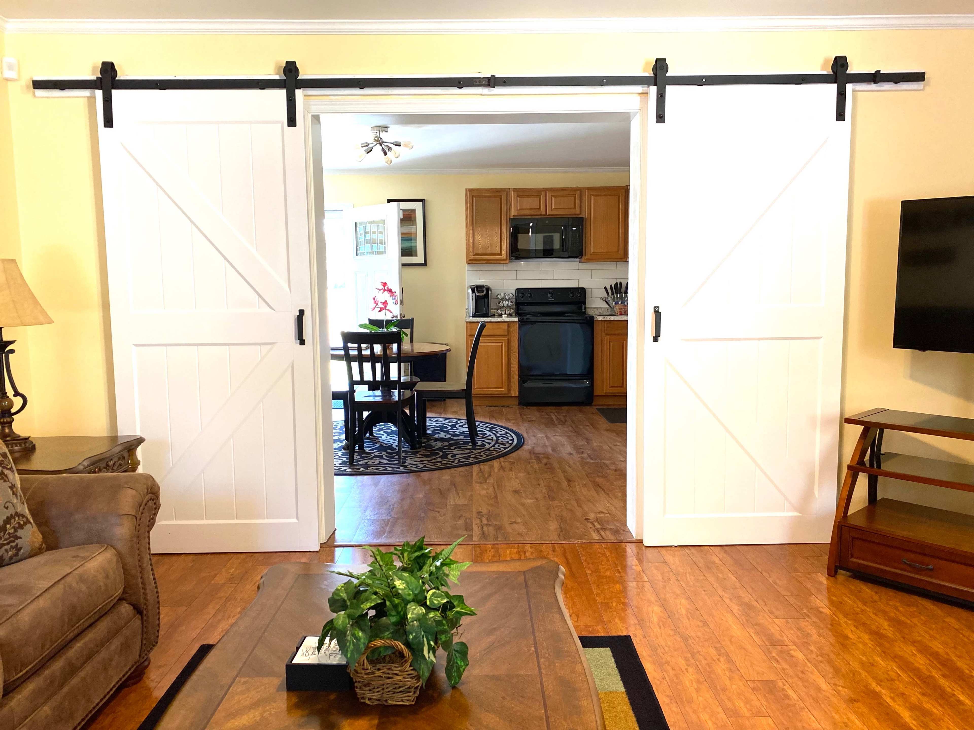 The image shows a living room with a large coffee table and a potted plant, leading into a kitchen area through two sliding white barn doors.