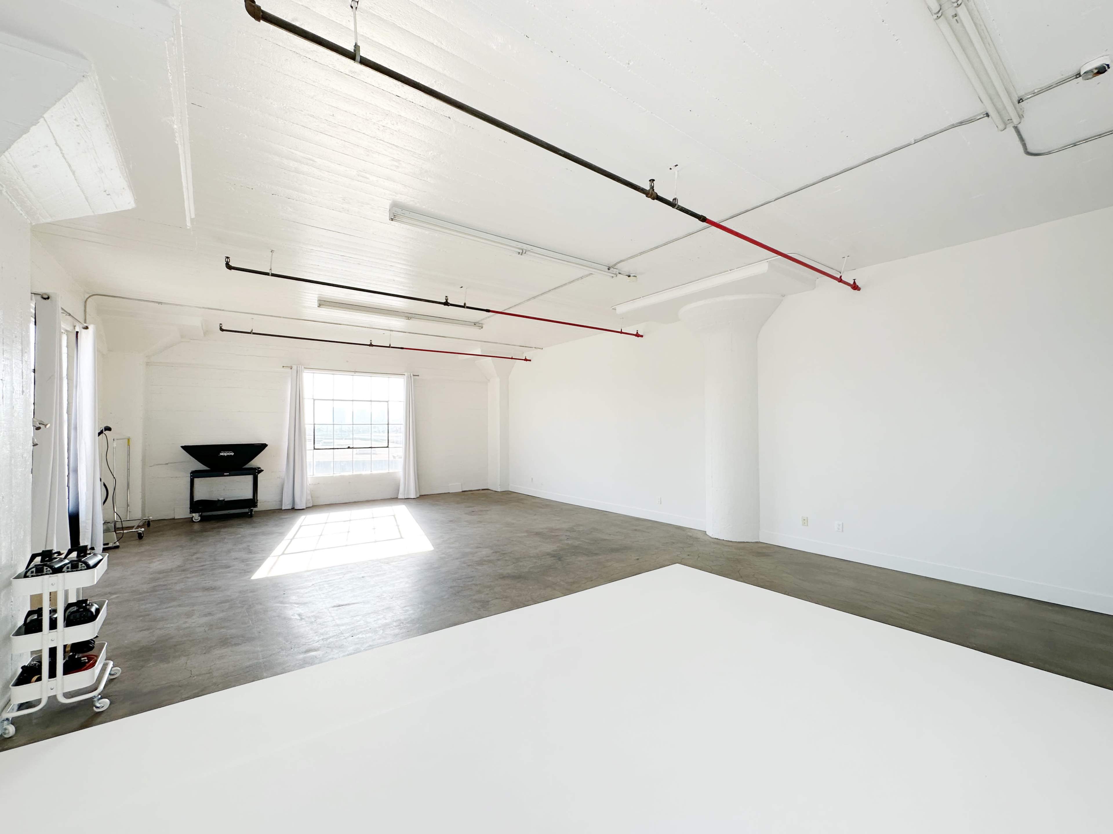 A spacious, empty room with white walls, a concrete floor, and large windows allowing natural light to enter.
