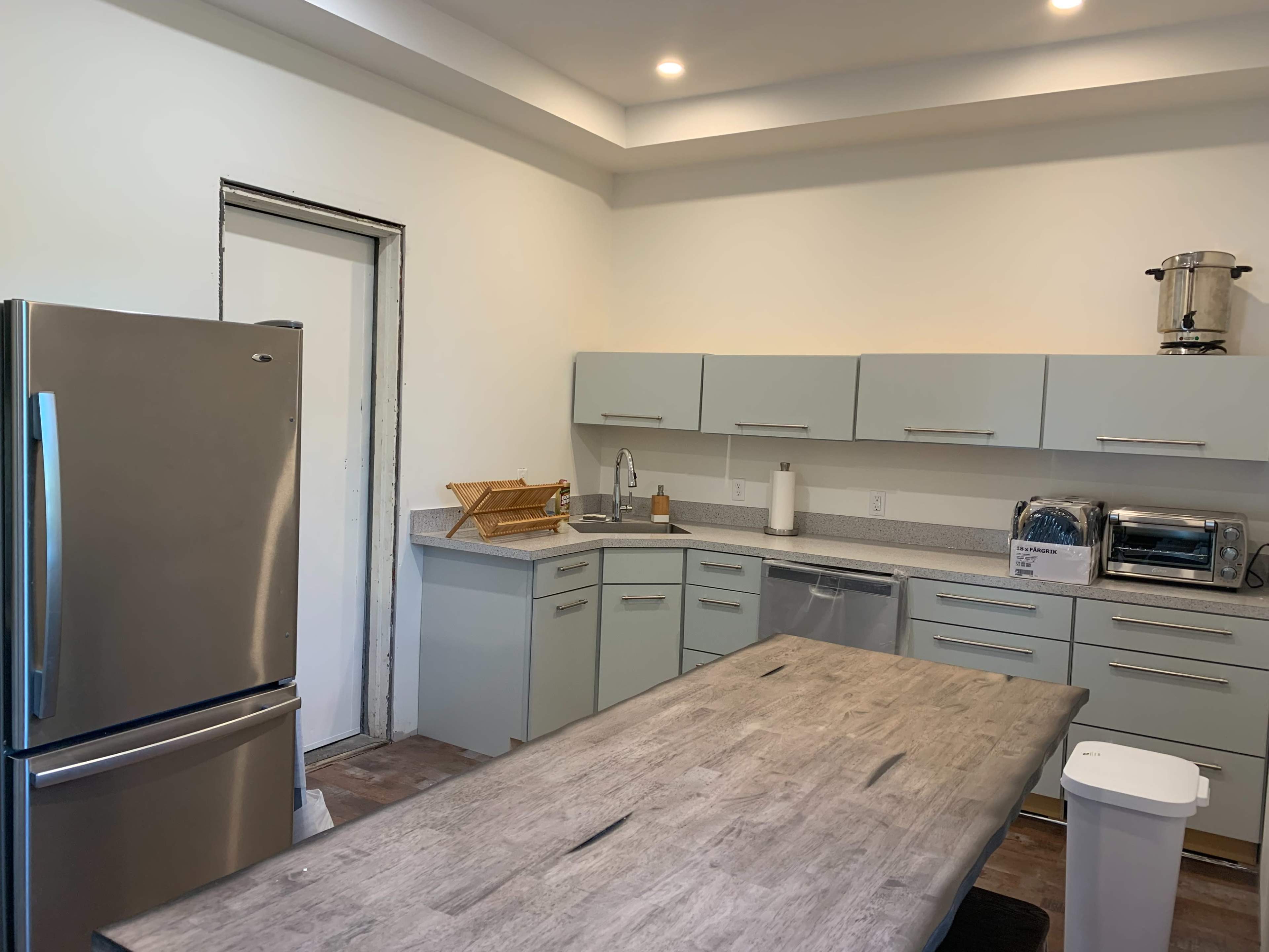 A modern kitchen features a large wooden table, stainless steel refrigerator, and light green cabinetry.