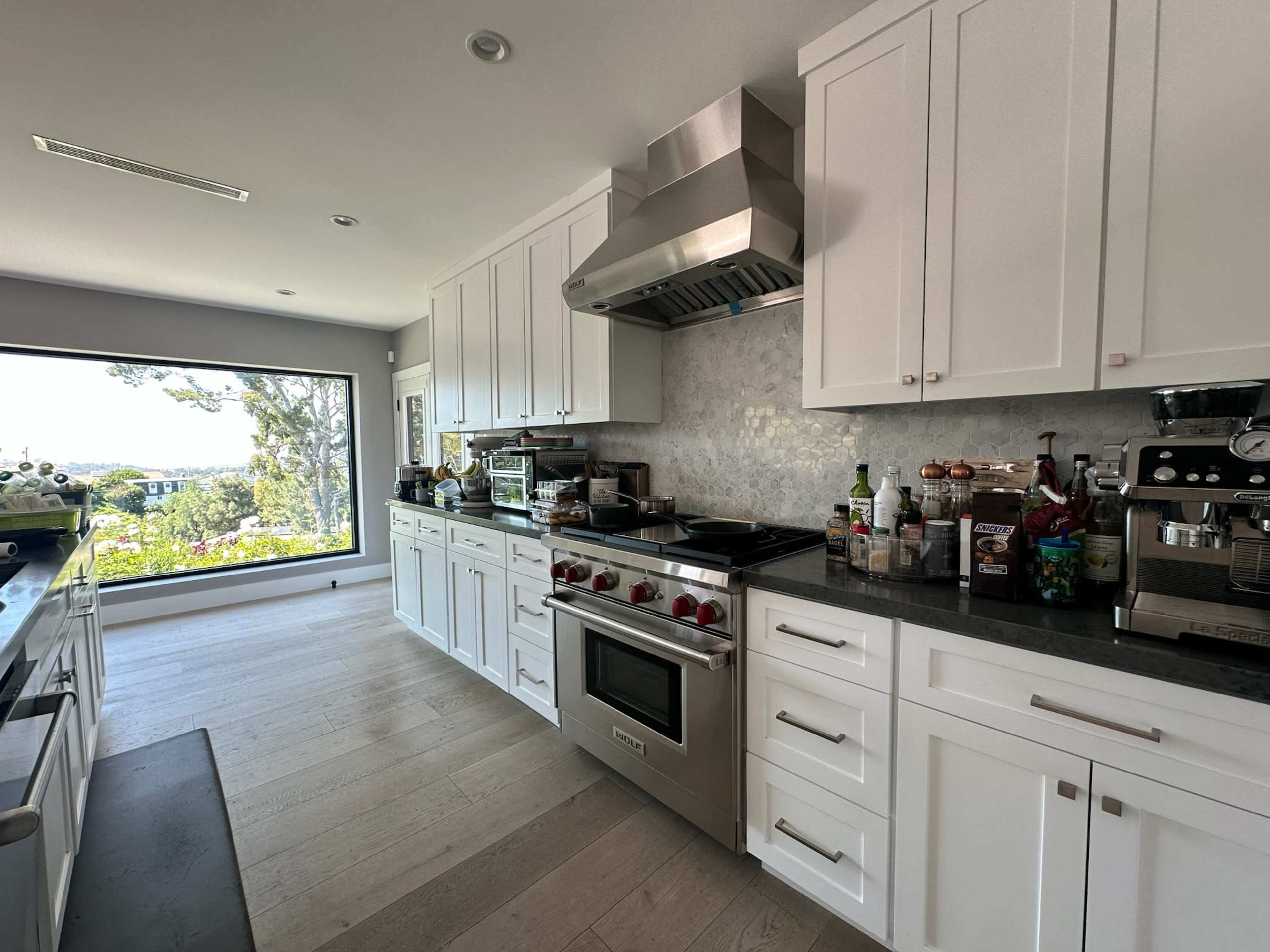 A modern kitchen features white cabinetry, a large window with a view, and stainless steel appliances, including a gas stove and coffee machine.