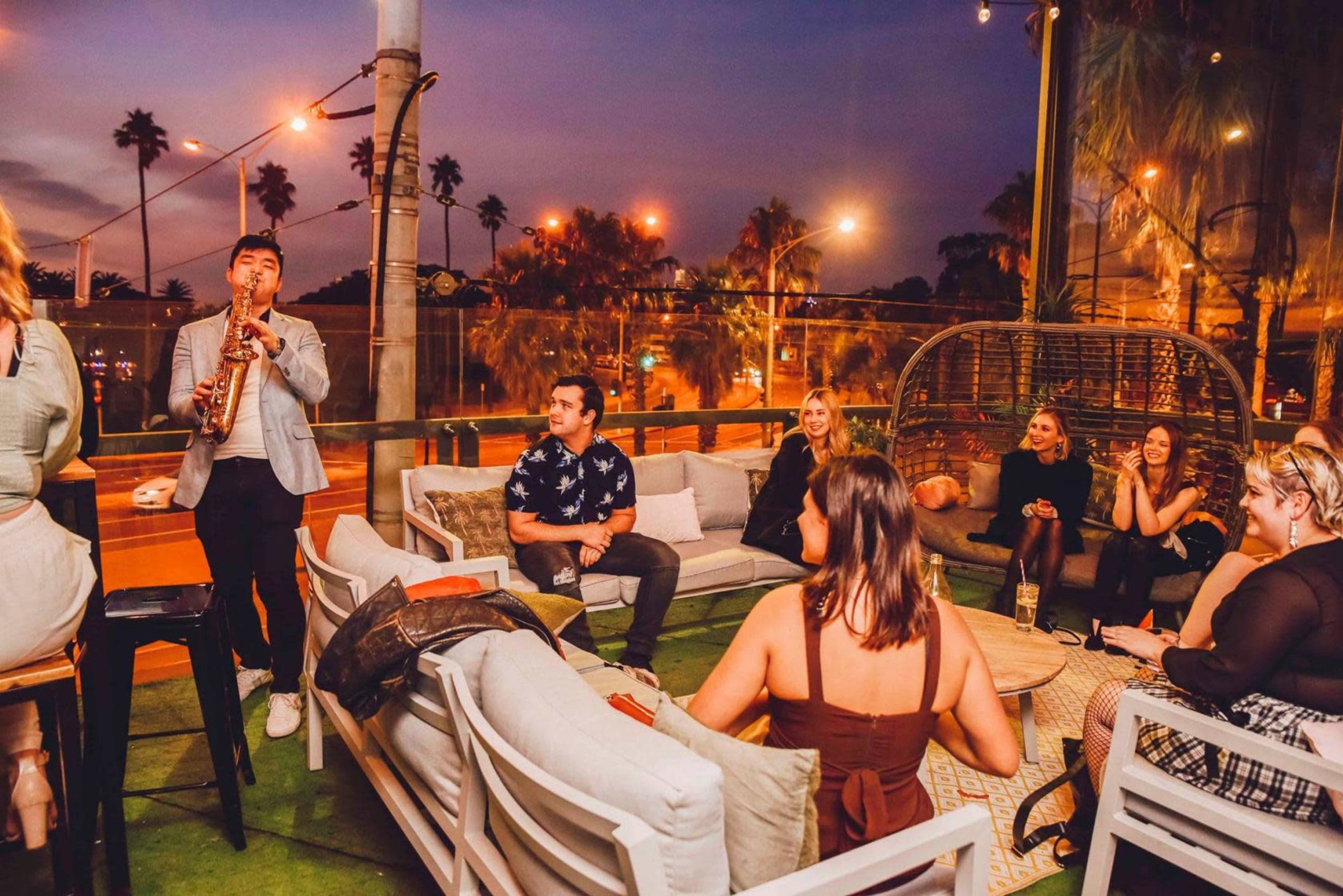 A musician plays the saxophone on a rooftop lounge, while several people socialize on comfortable seating in a dimly lit atmosphere.