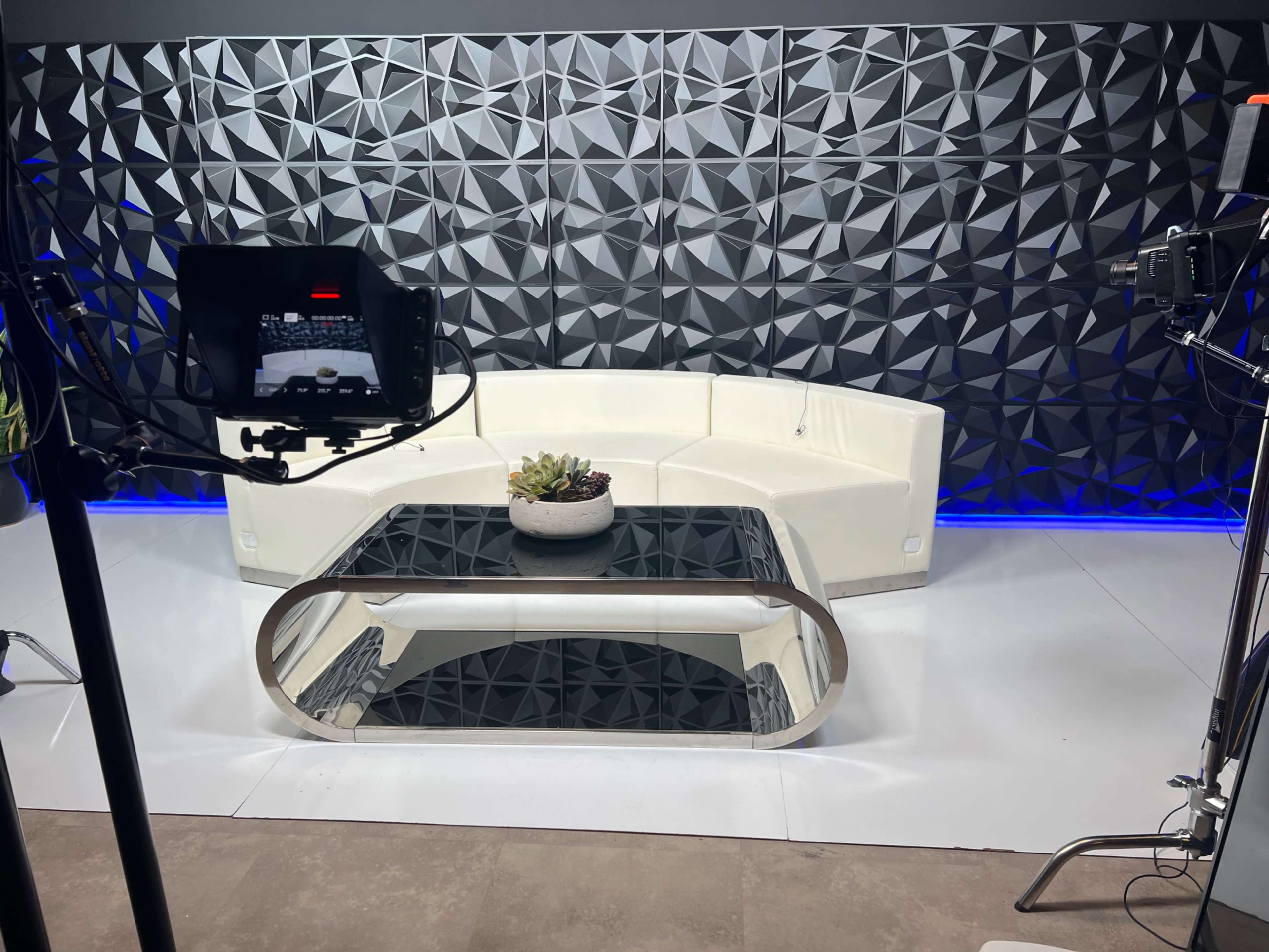 A modern studio setting with a white sectional sofa, a glass coffee table featuring a plant centerpiece, and a textured geometric wall backdrop illuminated by blue lighting.