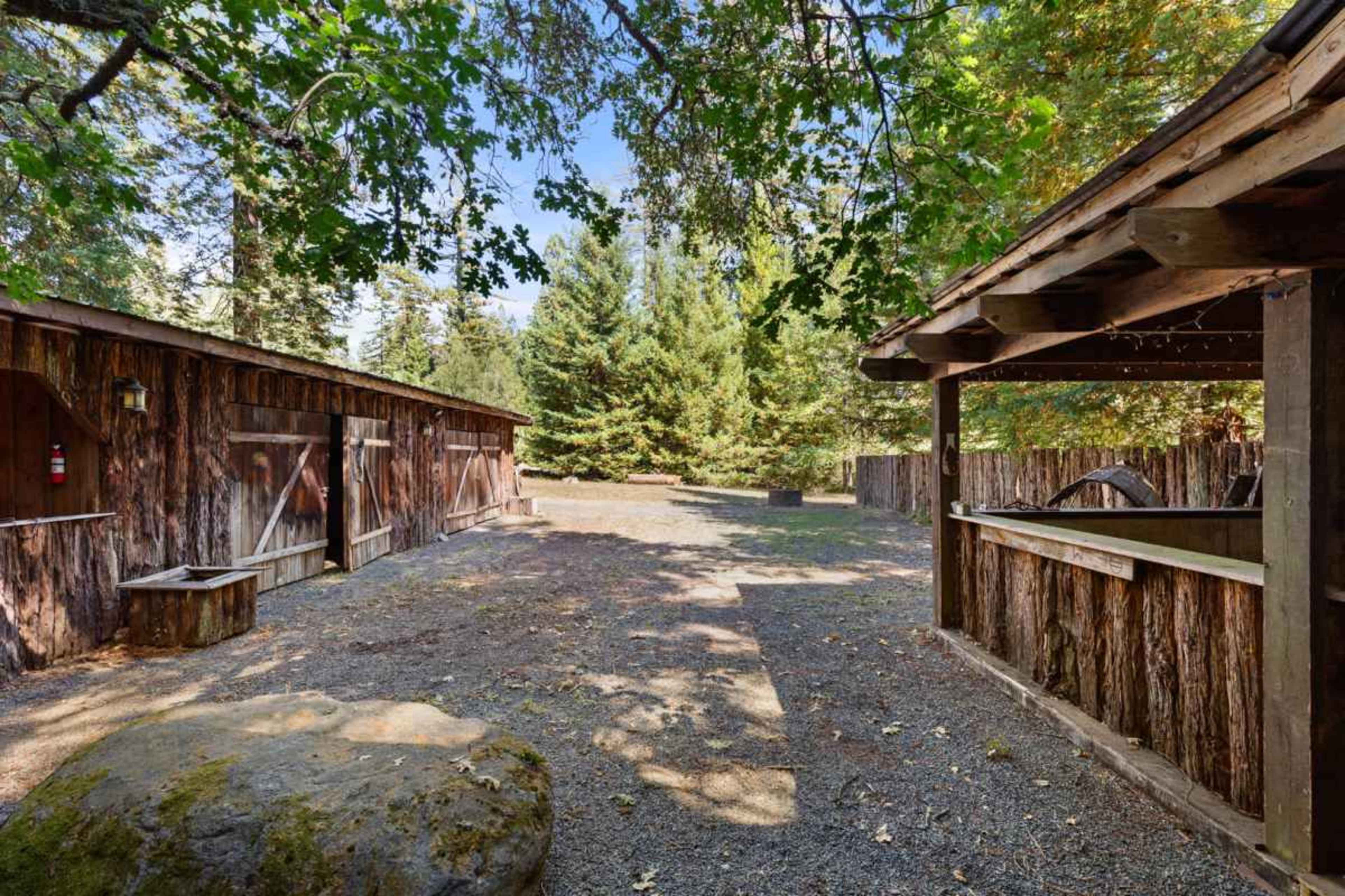 The image shows a rustic outdoor area with wooden structures and a gravel path surrounded by trees.