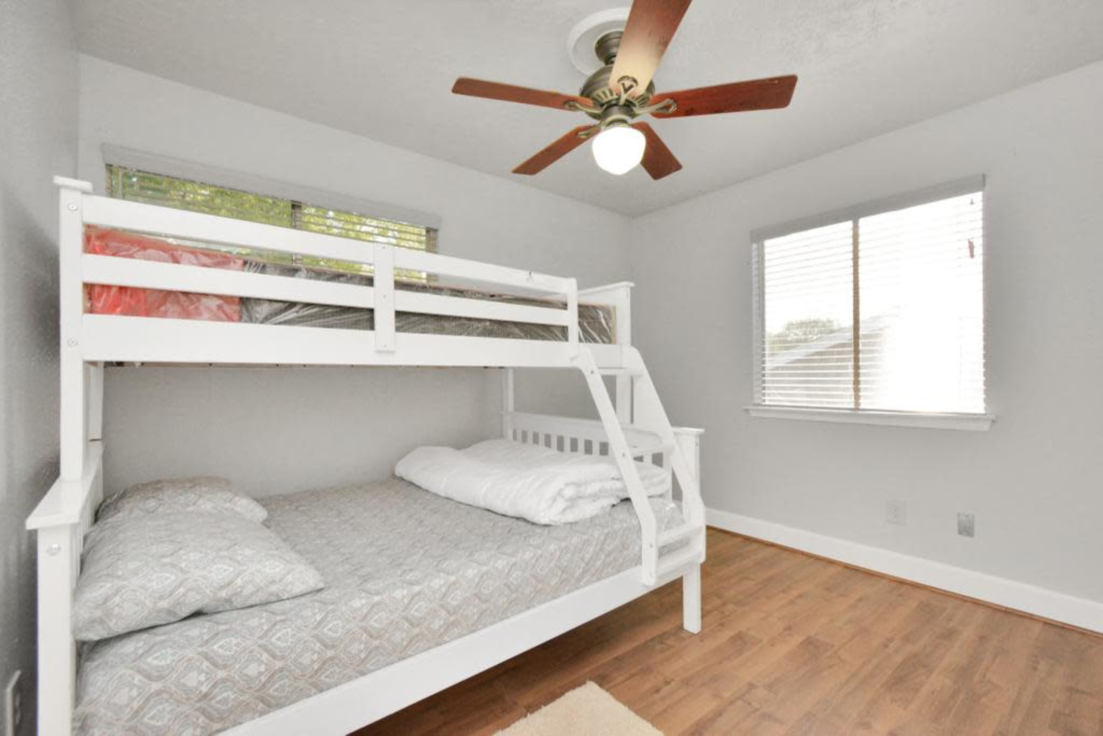 The image shows a bedroom featuring a white bunk bed, a single bed on top and a double bed below, with a ceiling fan and a window letting in natural light.
