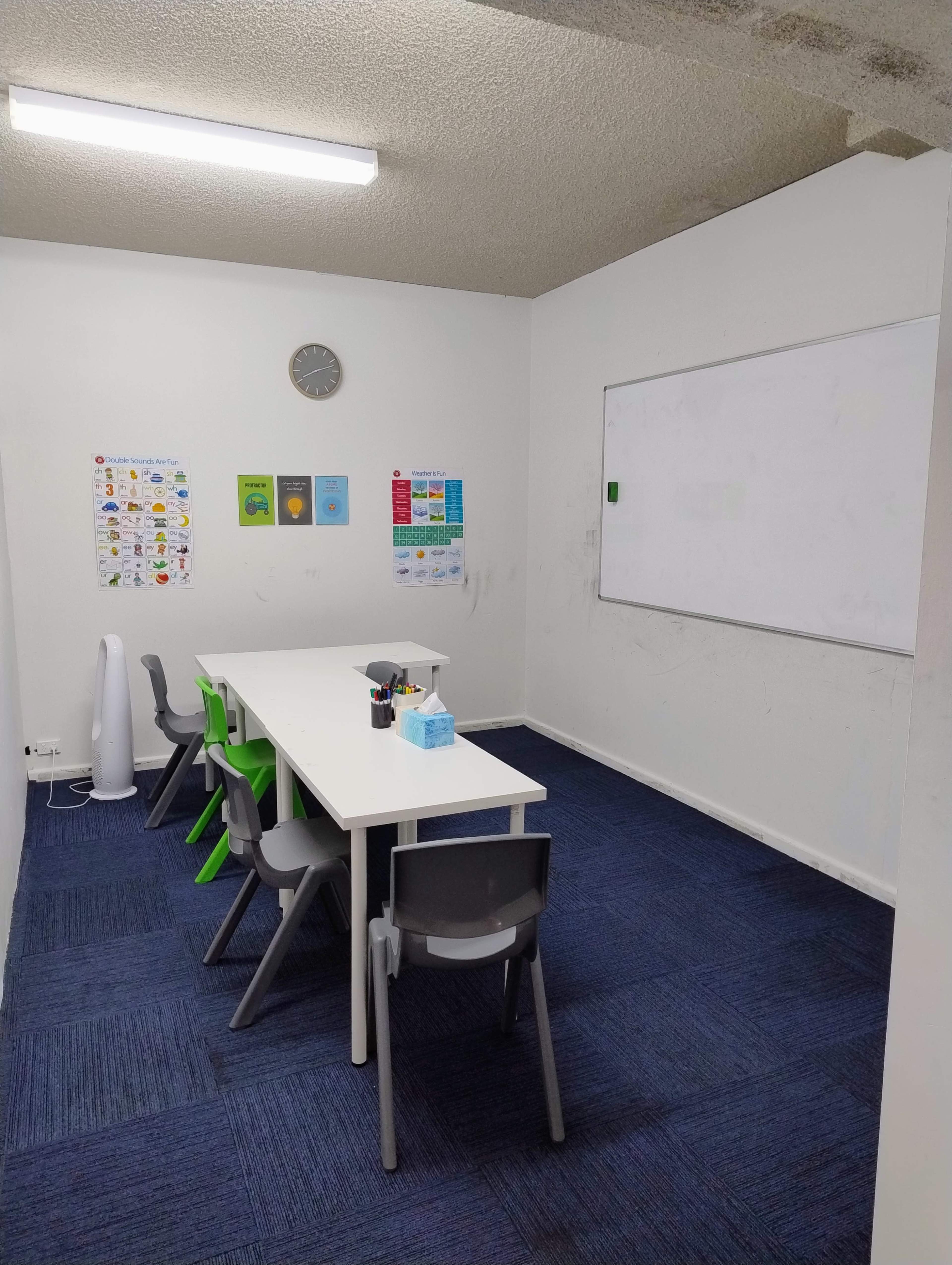 A small classroom features a white table surrounded by chairs, a whiteboard on the wall, and educational posters displayed.