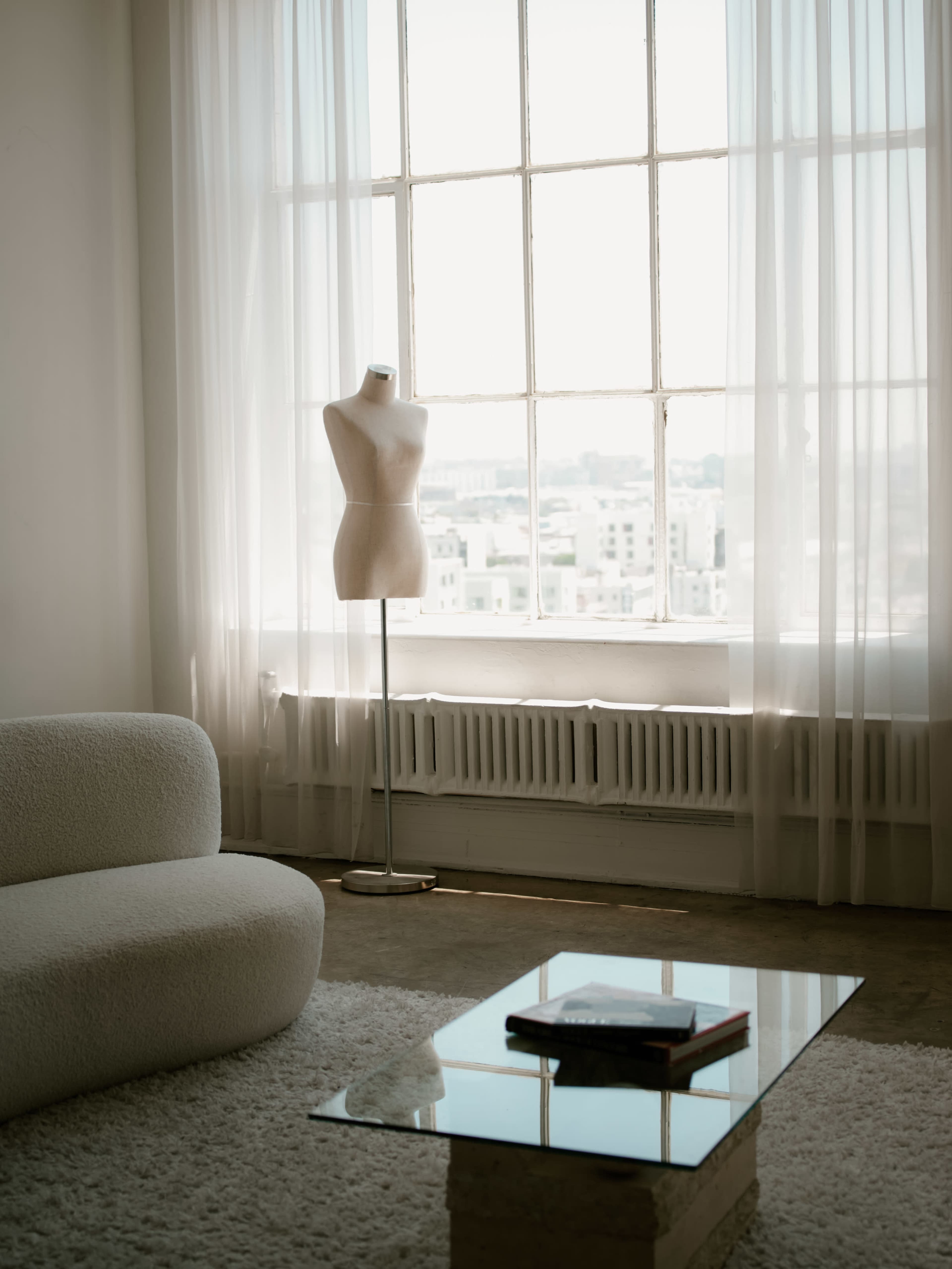 A mannequin stands beside a large window, with sheer curtains letting in natural light, and a glass coffee table sits on a soft rug.