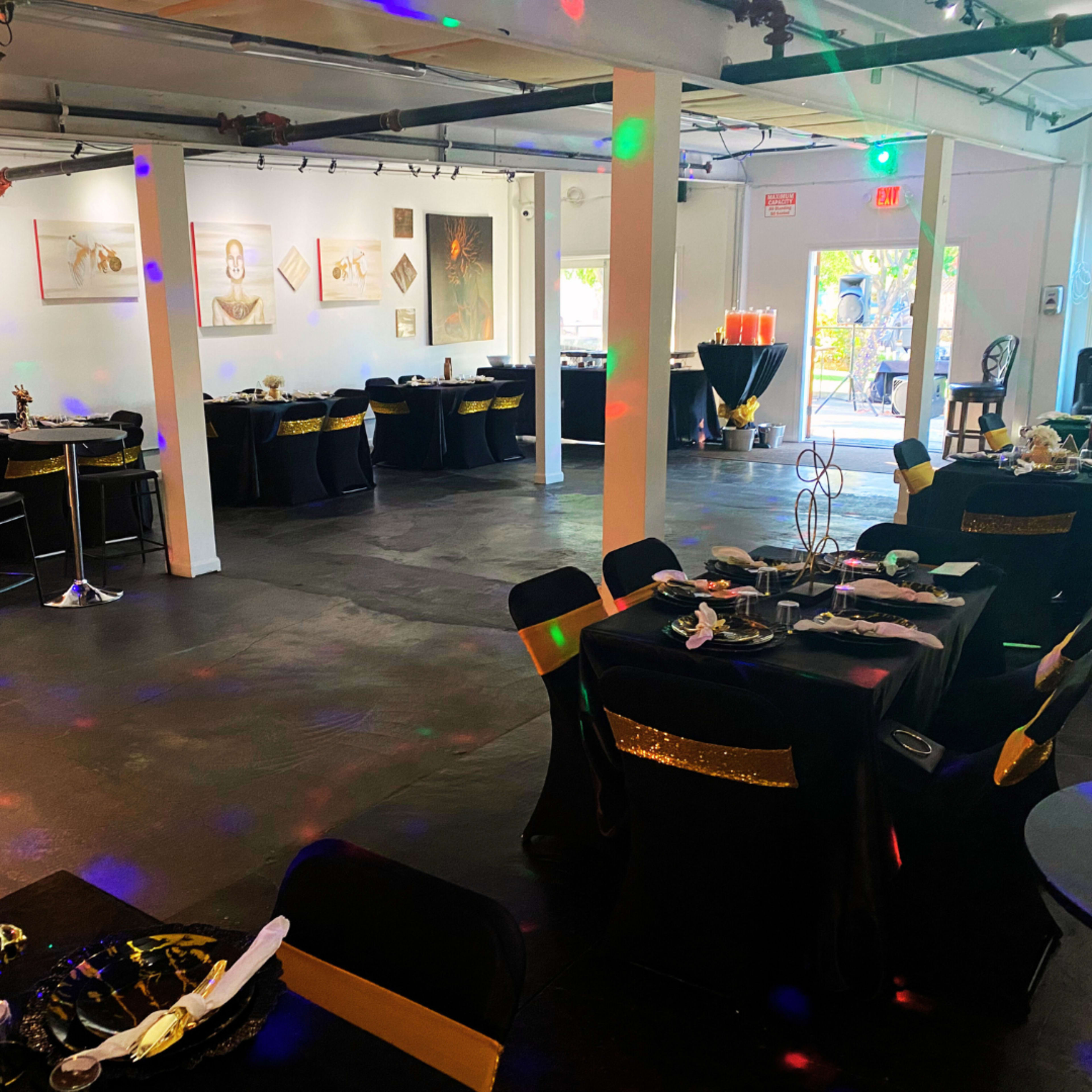 A decorated event space features tables with black and gold tablecloths, colorful lighting effects, and artwork on the walls.
