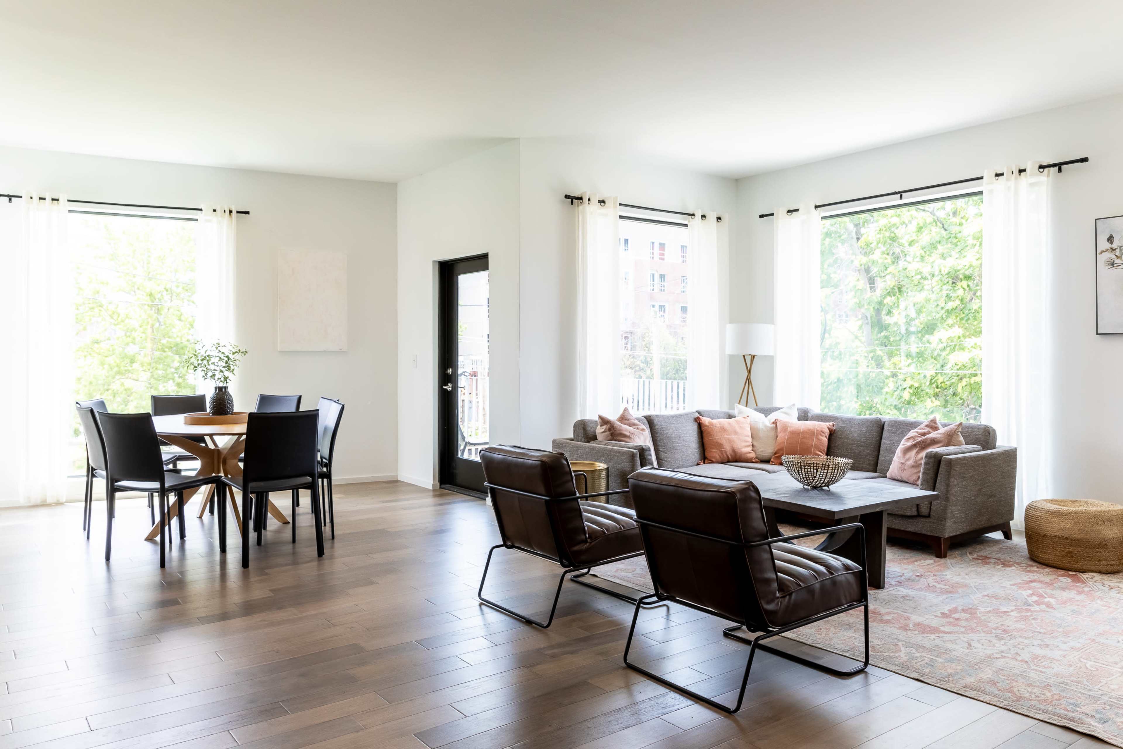 A bright, open living space featuring a seating area with a gray sofa and black chairs, a dining table for four, and large windows letting in natural light.