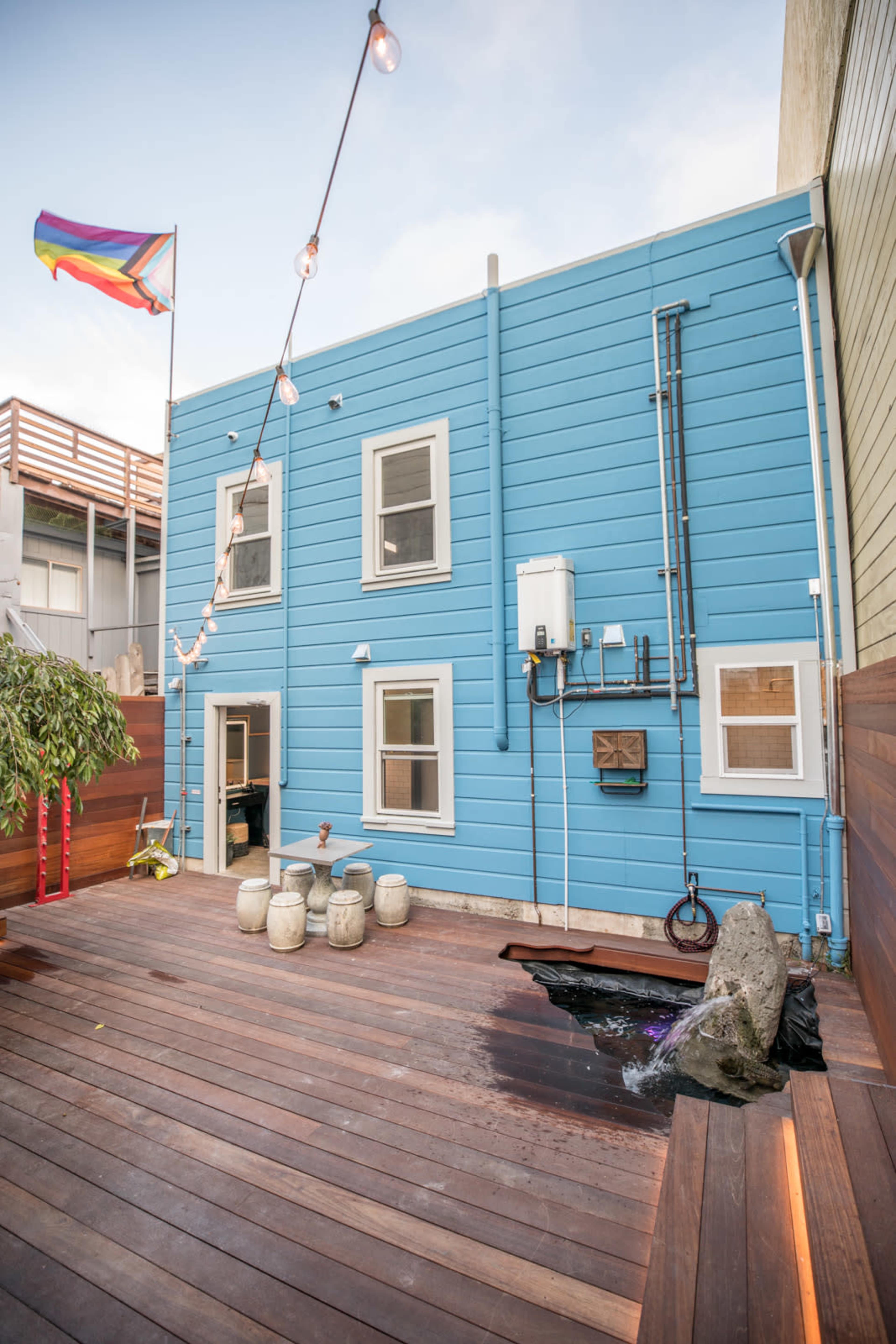 The image shows a blue wooden house with multiple windows and a colorful flag flying above, accompanied by a wooden deck that features a small water feature and decorative stone elements.