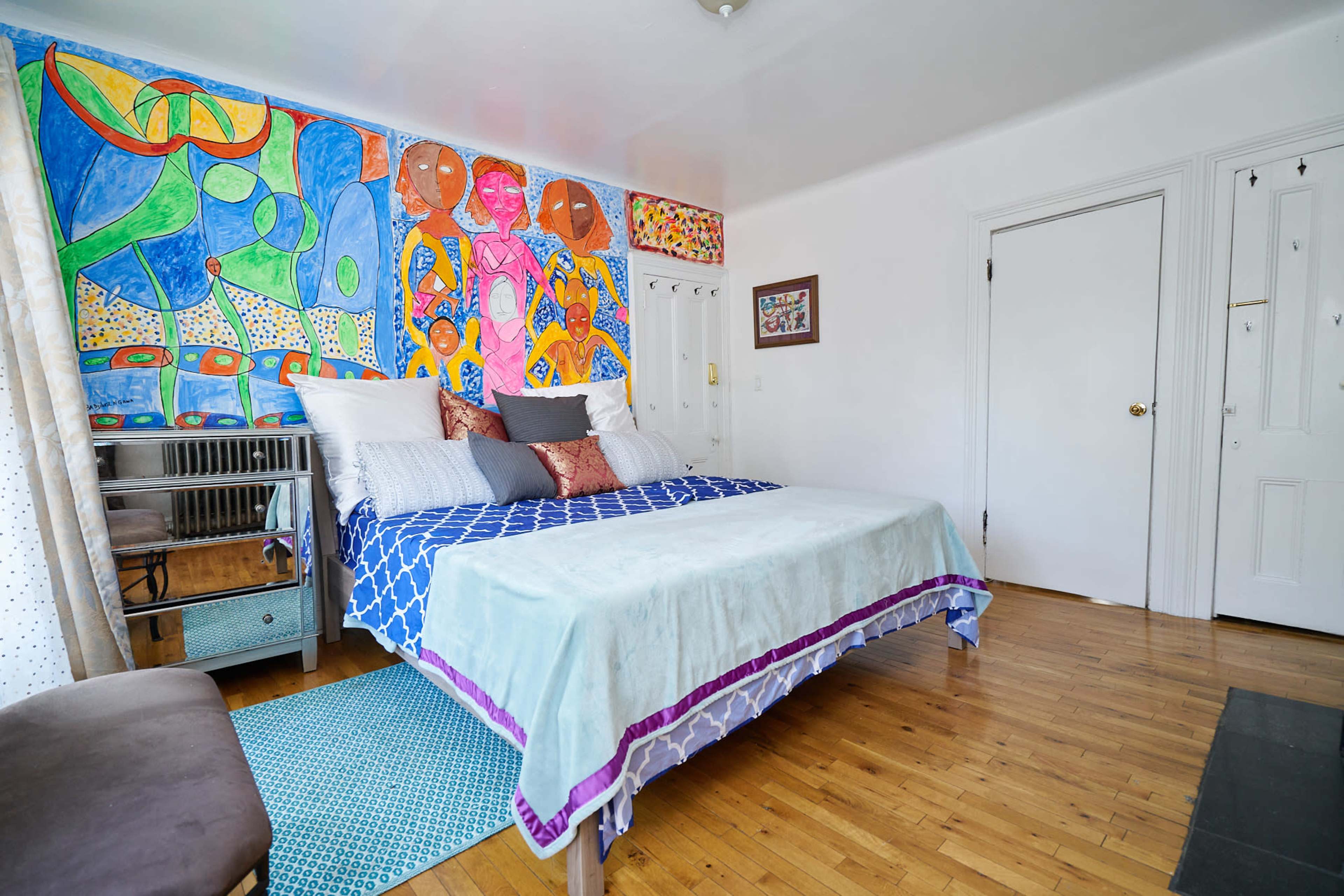 The image shows a bedroom with a colorful mural on one wall, featuring a large bed with decorative pillows and wooden flooring.