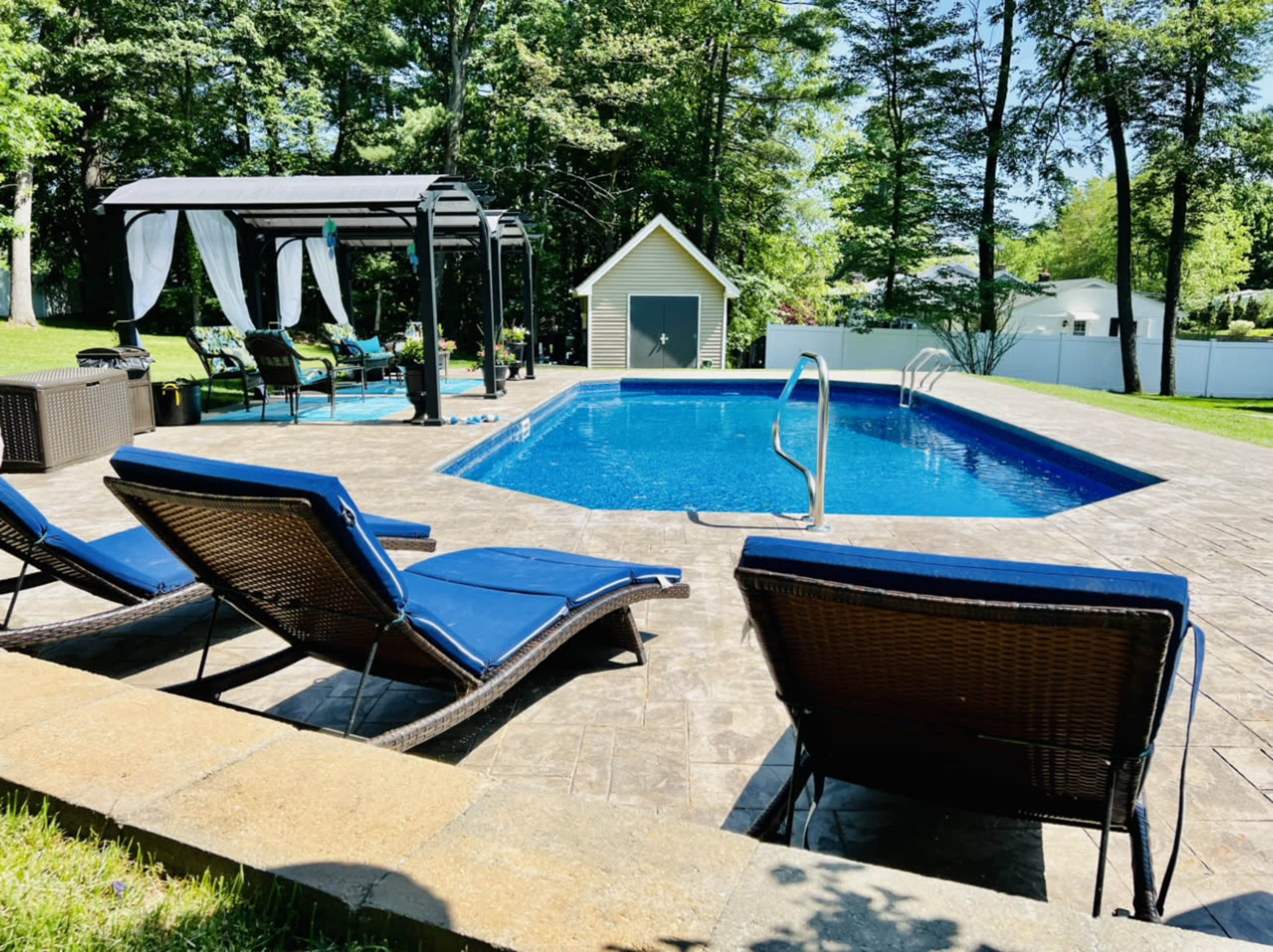 The image shows a backyard swimming pool surrounded by lounge chairs, a gazebo, and a shed, with trees in the background.