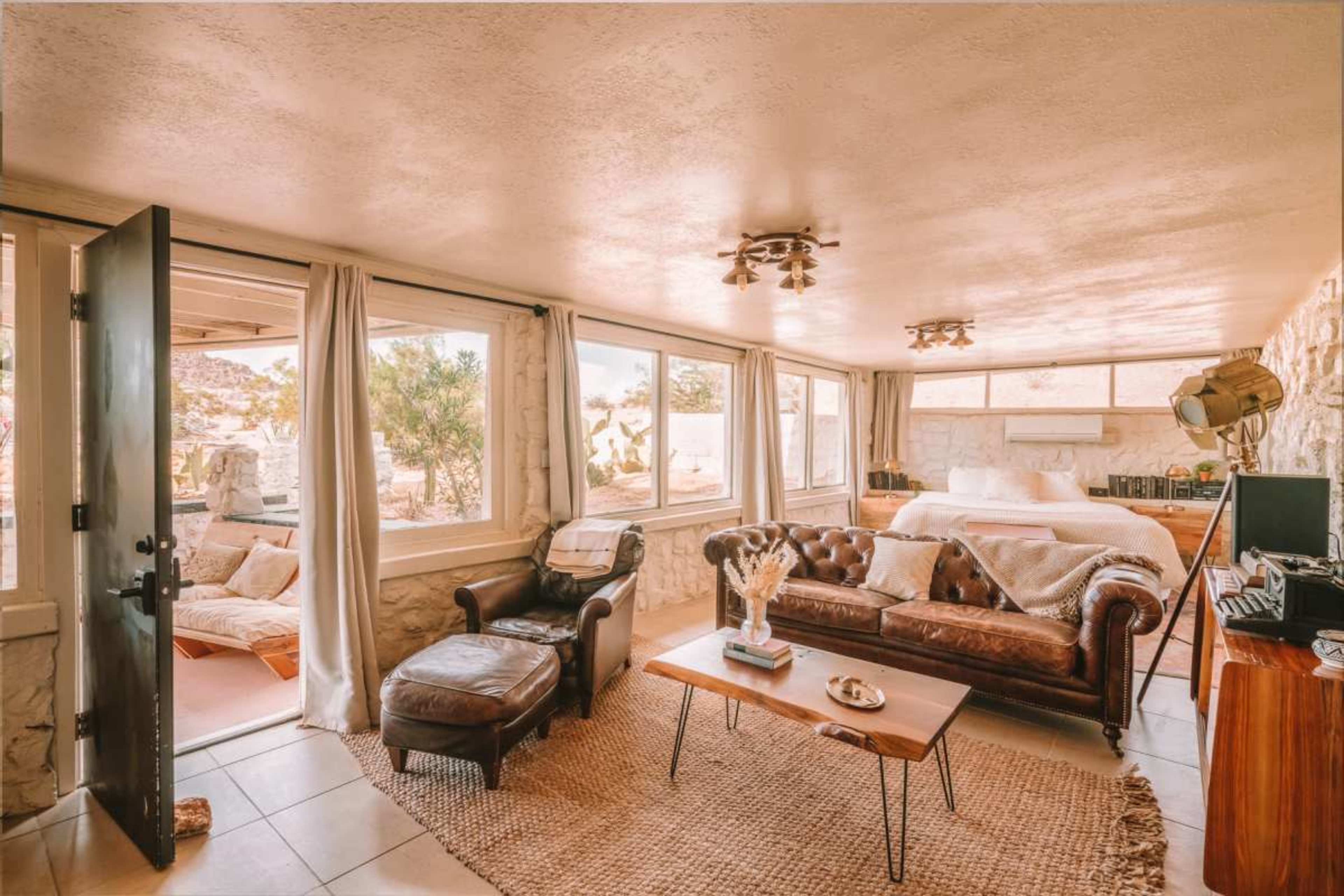 The interior of a cozy living space features a leather sofa, a coffee table, and large windows that offer views of desert landscaping.